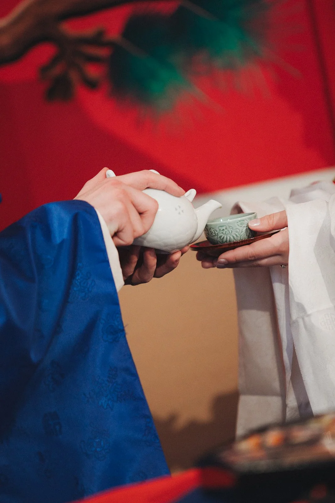 A person in a white robe is receiving tea from another person wearing a blue garment, in an indoor setting with a red wall and artwork in the background.