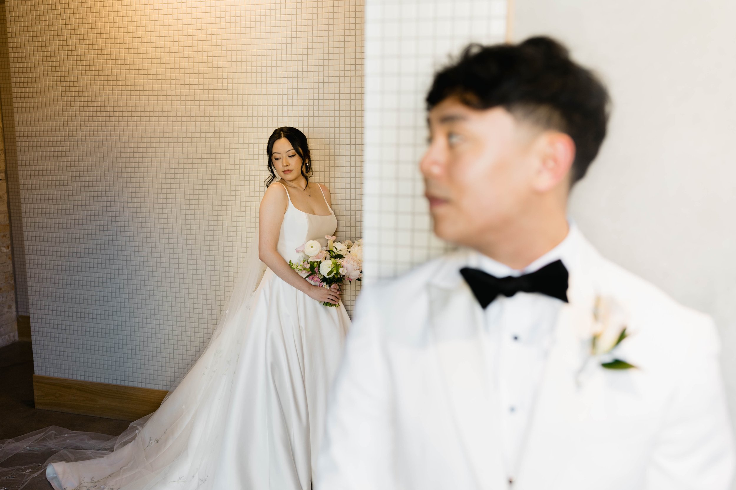 A bride in a white wedding dress holding a bouquet of flowers, and a groom in a tuxedo with a bow tie, standing against a tiled wall backdrop.