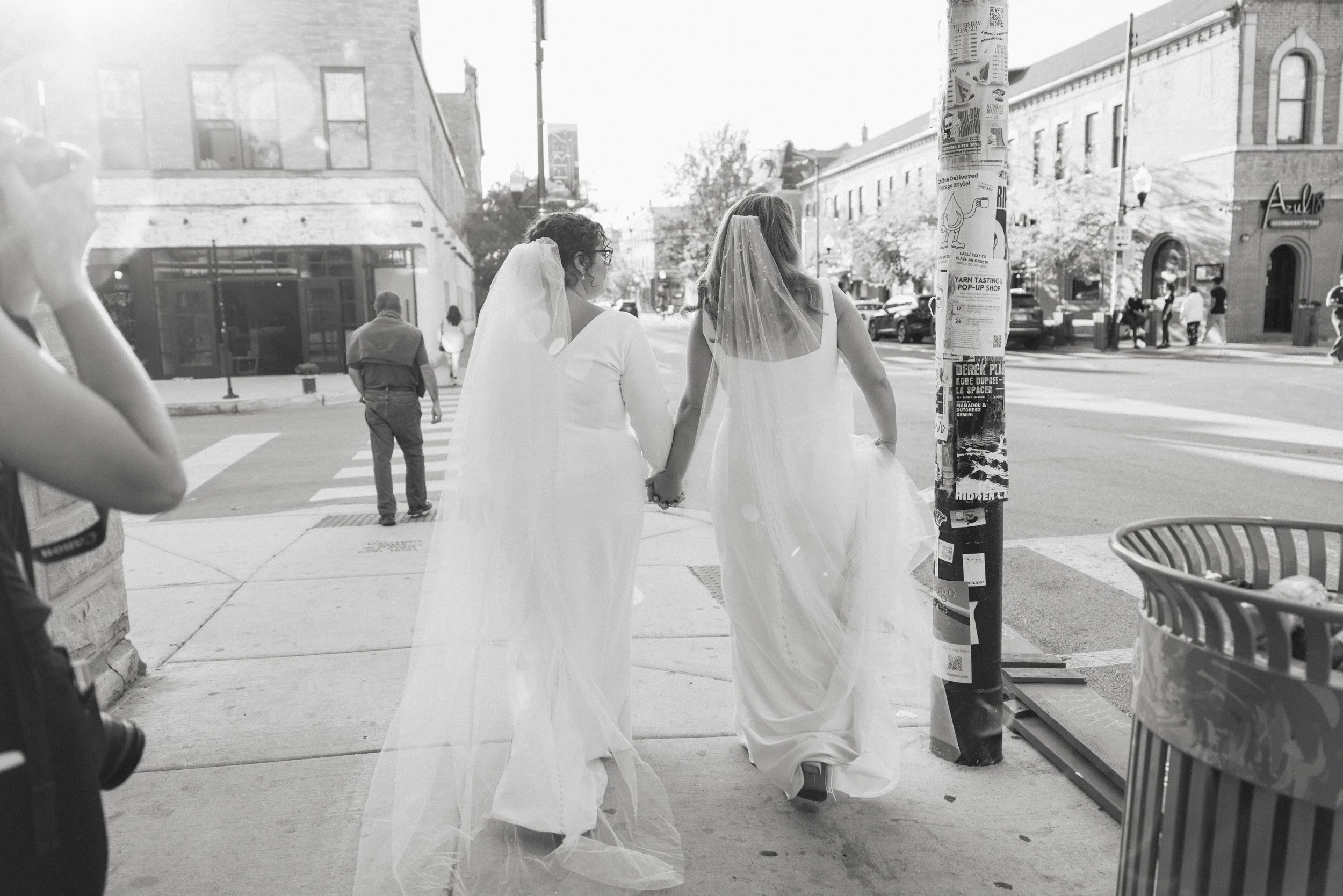 Two women in wedding dresses holding hands and walking on a city sidewalk, with other pedestrians and storefronts visible in the background.