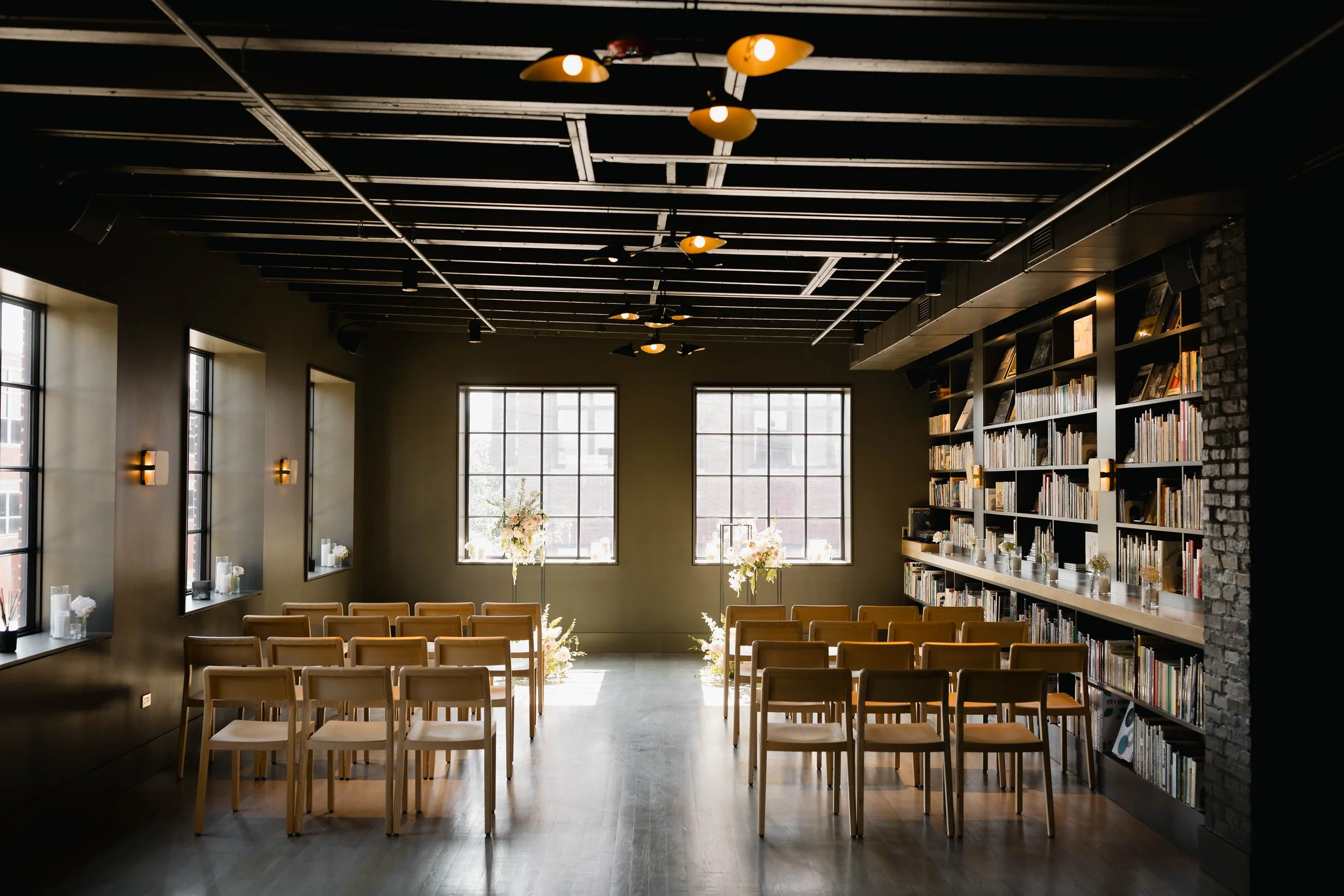 Interior of a modern library or event space with large windows, wooden chairs, many books on shelves, and decorative flowers.