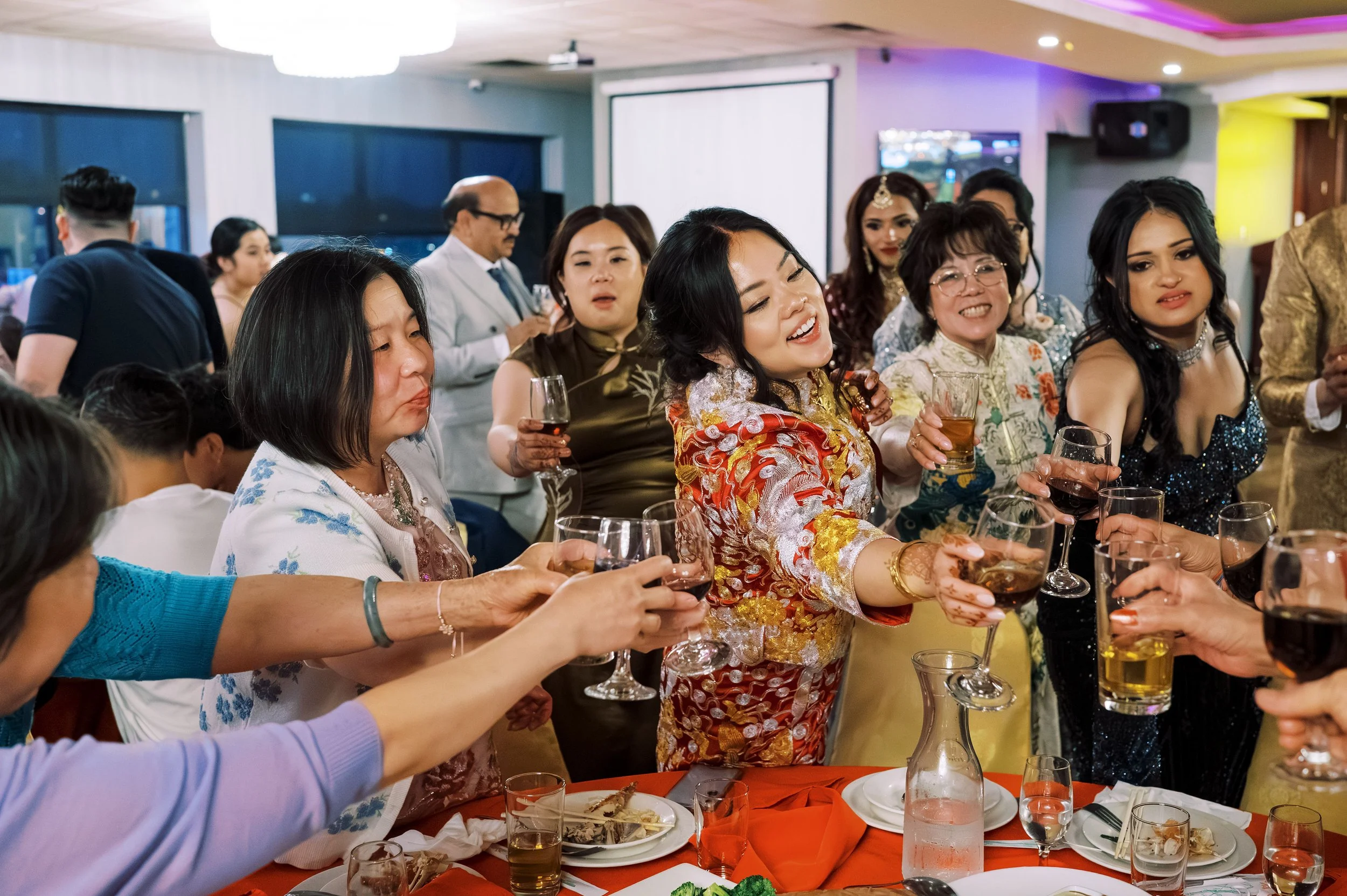People celebrating at a banquet table with drinks and plates, wearing colorful traditional attire, in a decorated event space.