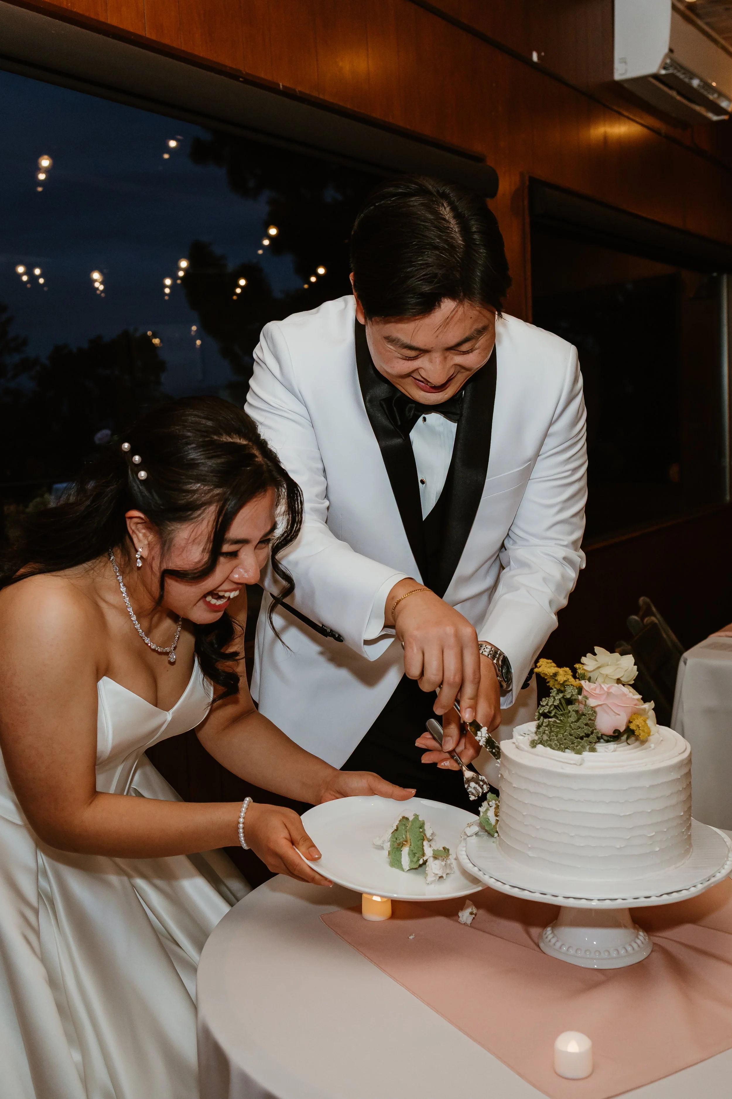 A bride and groom in wedding attire cutting a wedding cake together at their reception. The bride is in a white strapless gown with jewelry, and the groom is in a white tuxedo jacket with black lapels, a black bow tie, and a watch. There are floral d