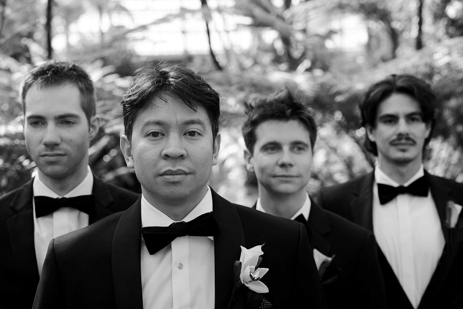 Black and white photo of four men in tuxedos with bow ties, standing outdoors with trees in the background. The man in the front center has a flower boutonniere on his lapel.