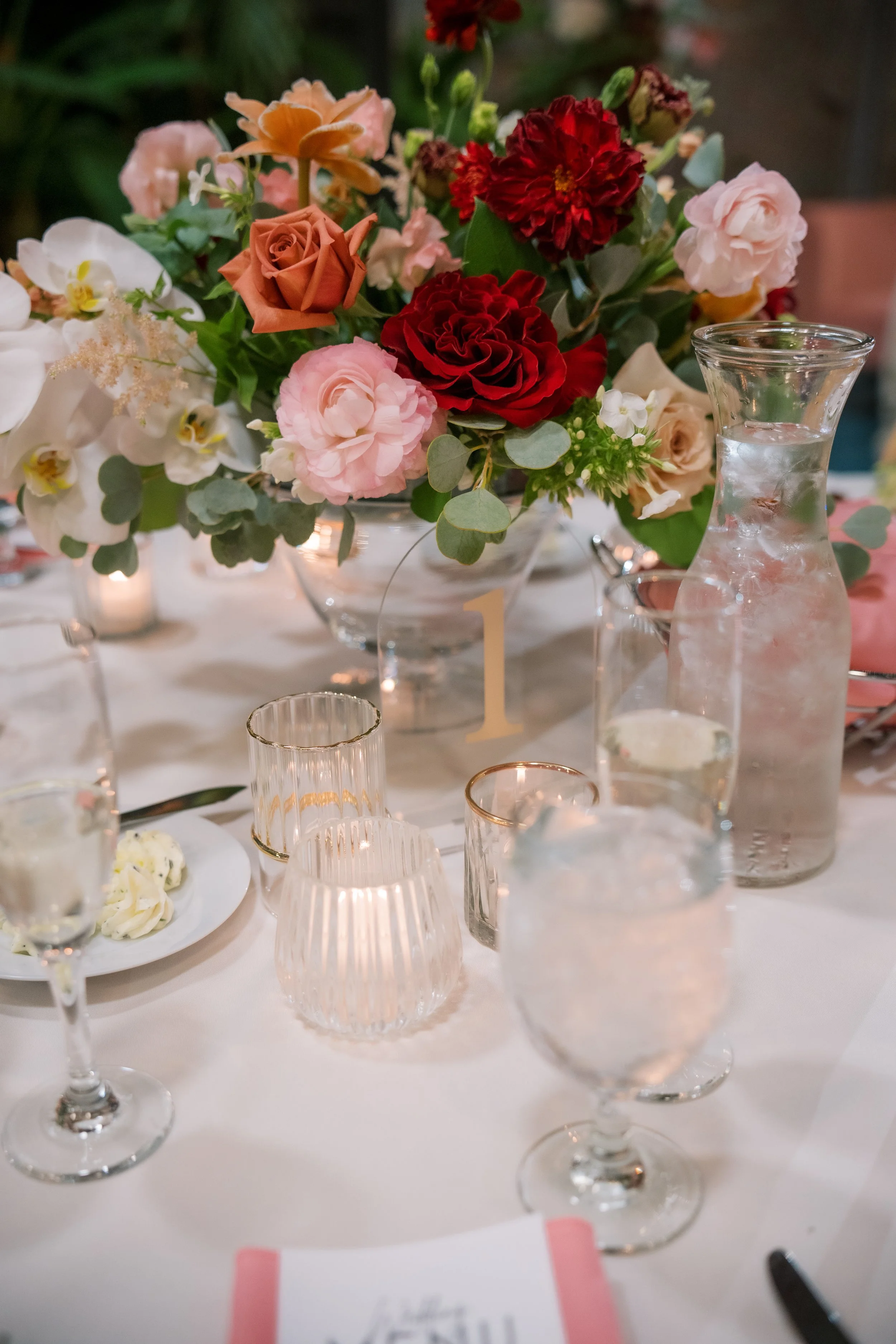 A floral centerpiece with pink, red, orange, and peach roses, along with white orchids, placed on a white tablecloth at a formal event. Glassware and candles are also visible on the table.