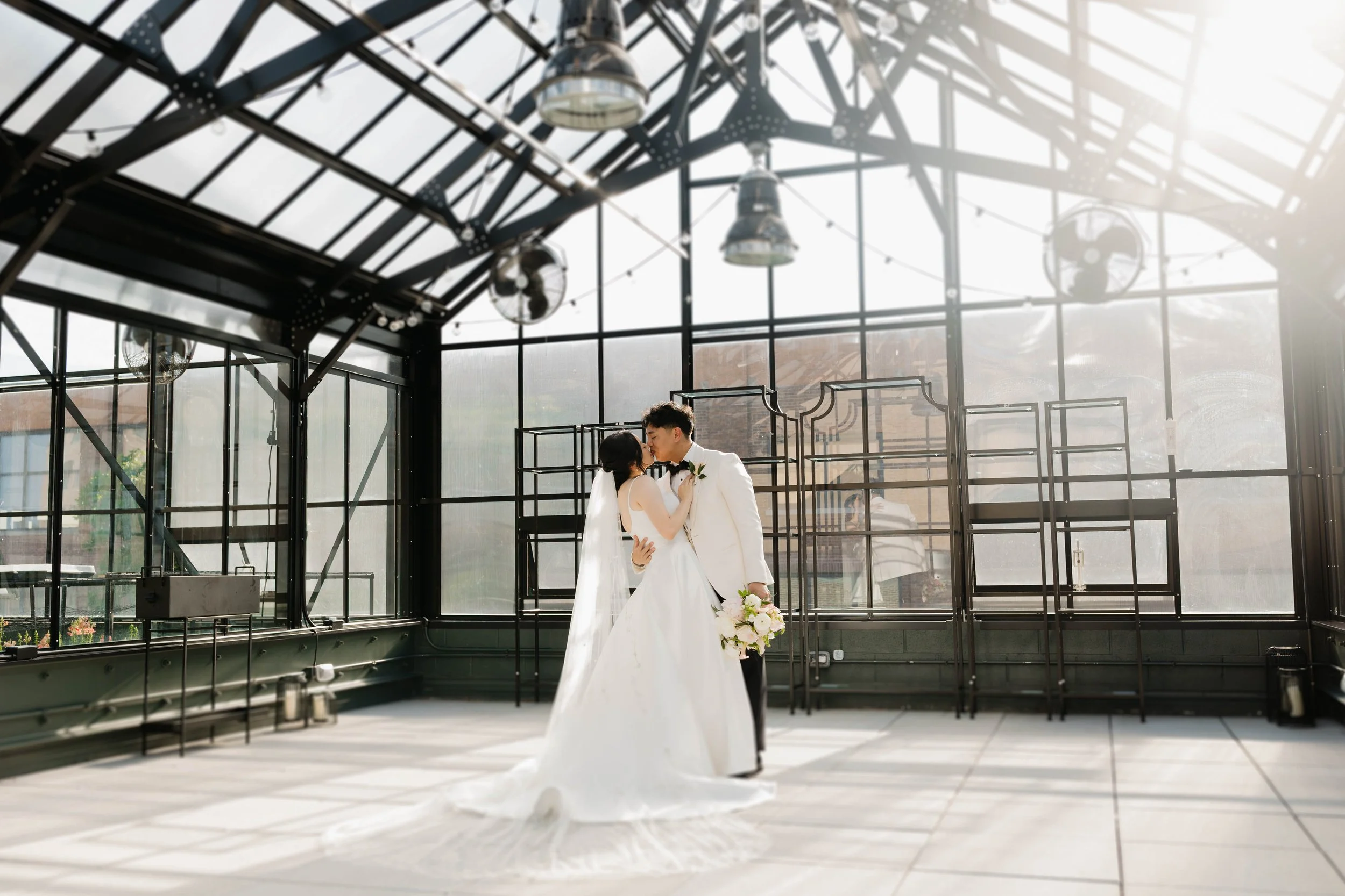 A bride and groom share a kiss inside a glass-walled industrial-style venue with metal beams and hanging lights, sunlight streaming through the windows.