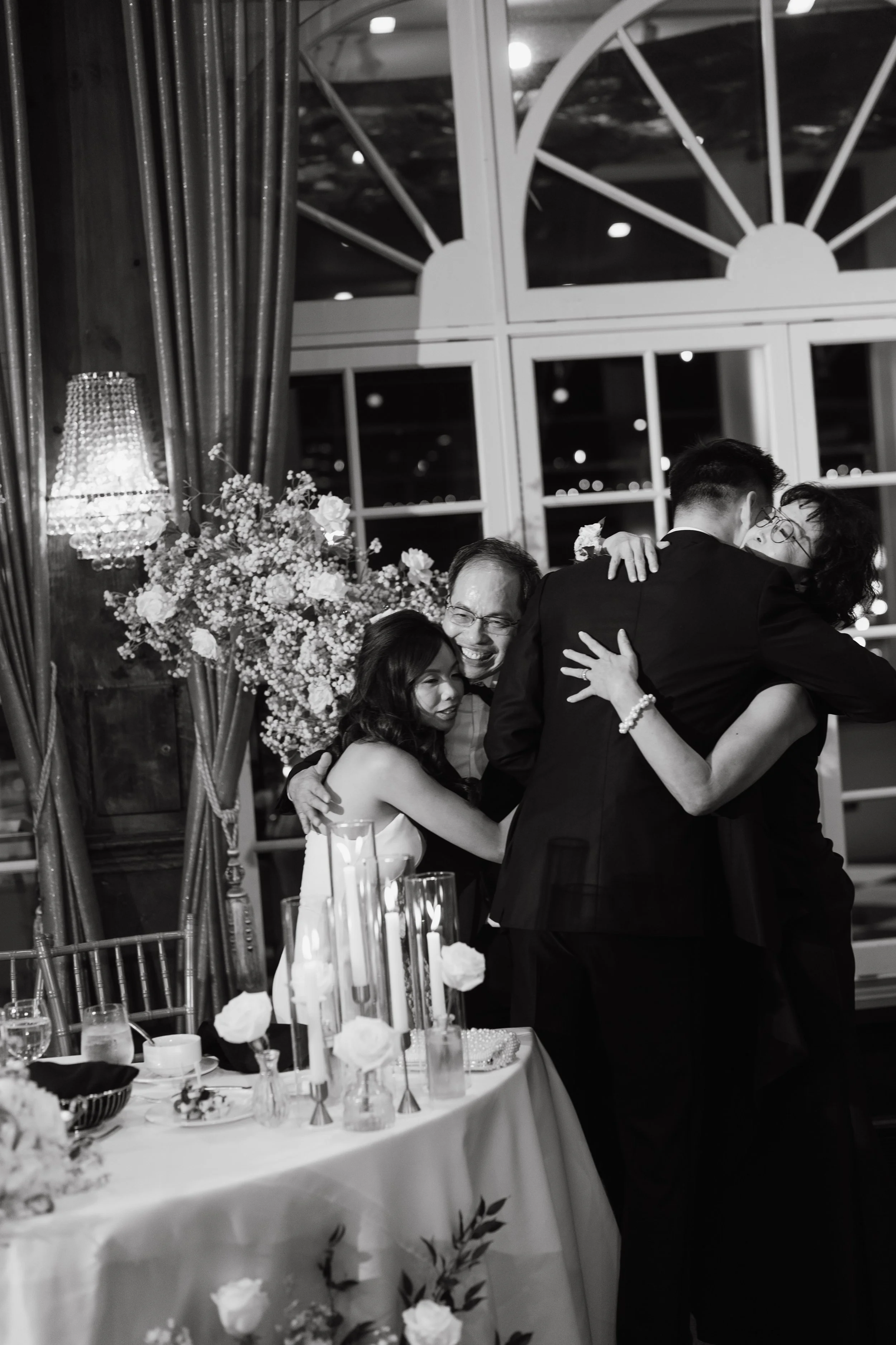 A black and white photo of a wedding reception with a group of people hugging and dancing near a table with floral arrangements and candles.