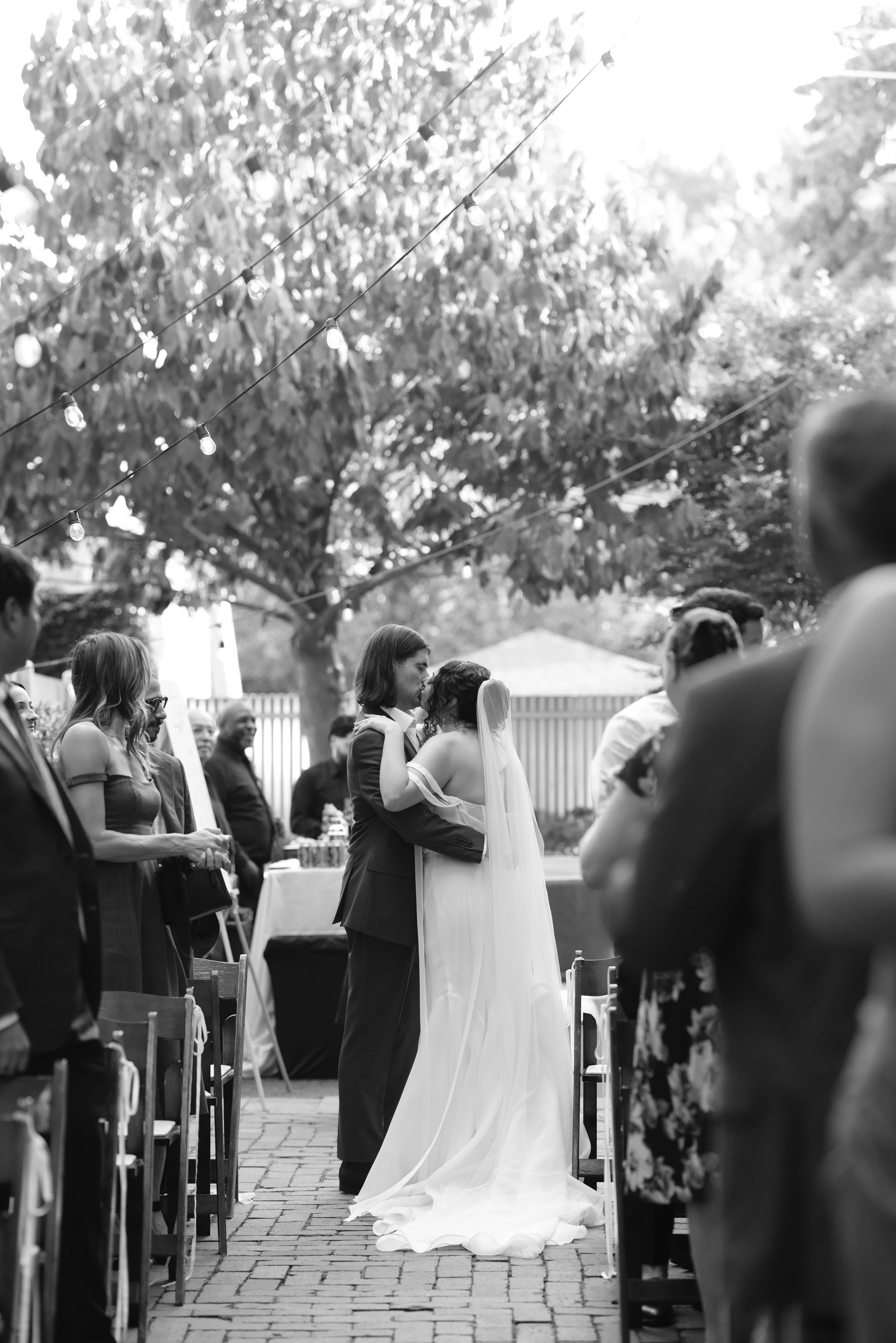 A bride and groom share a dance at their outdoor wedding ceremony, surrounded by seated guests. String lights are hanging overhead, and trees and a fence are in the background.