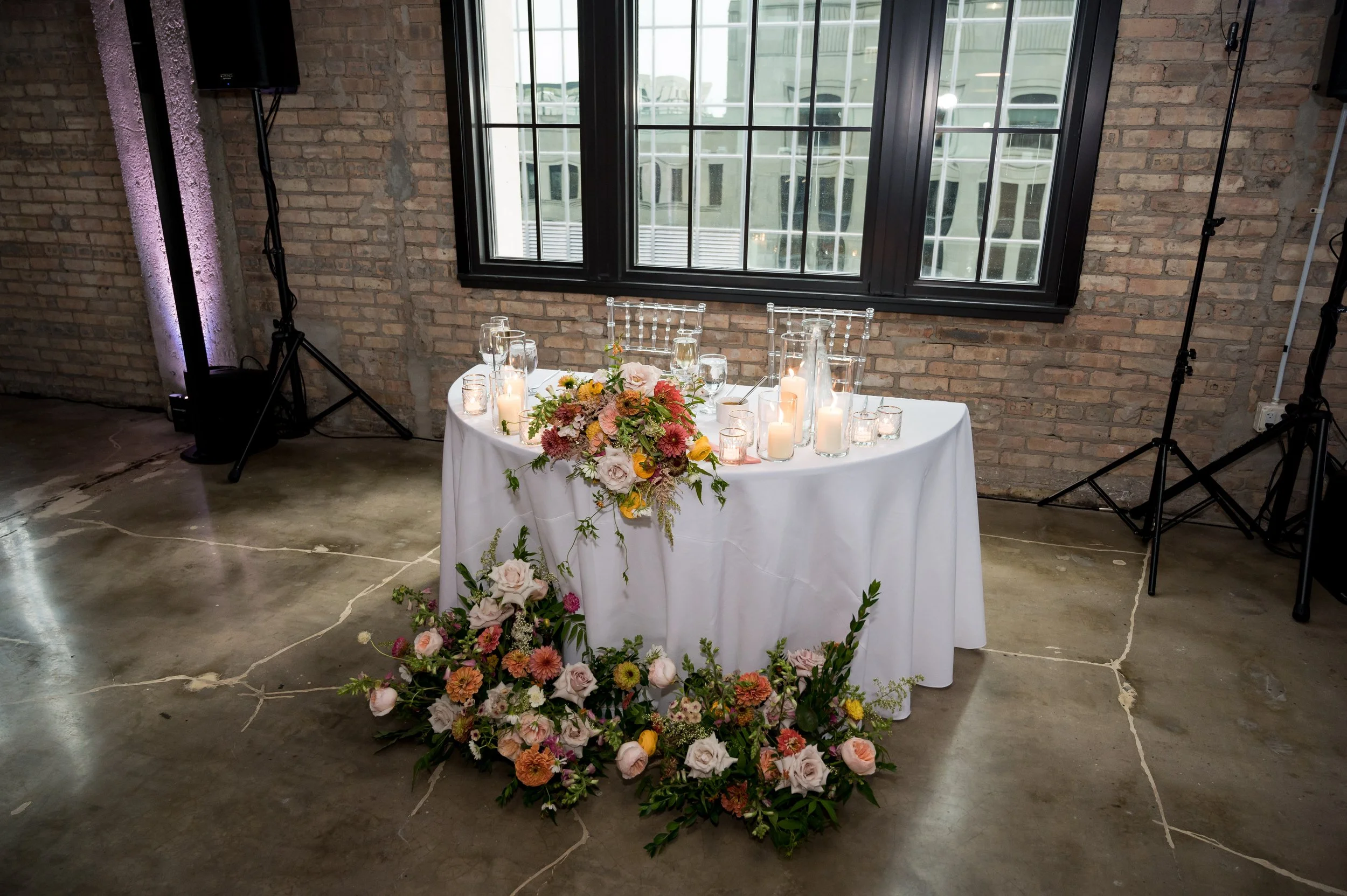 A table decorated with a floral centerpiece, surrounded by candles and glasses, set against a brick wall with large windows, in an indoor venue.