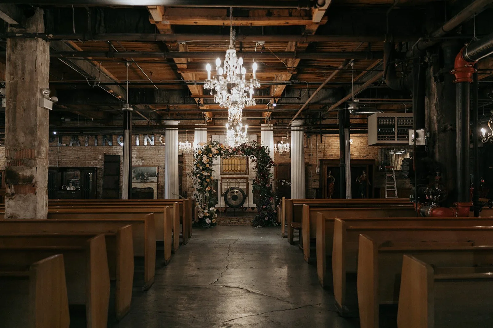 Interior of a rustic wedding venue with wooden pews, chandelier lighting, and a floral arch at the altar, exposed brick walls, and industrial pipes.