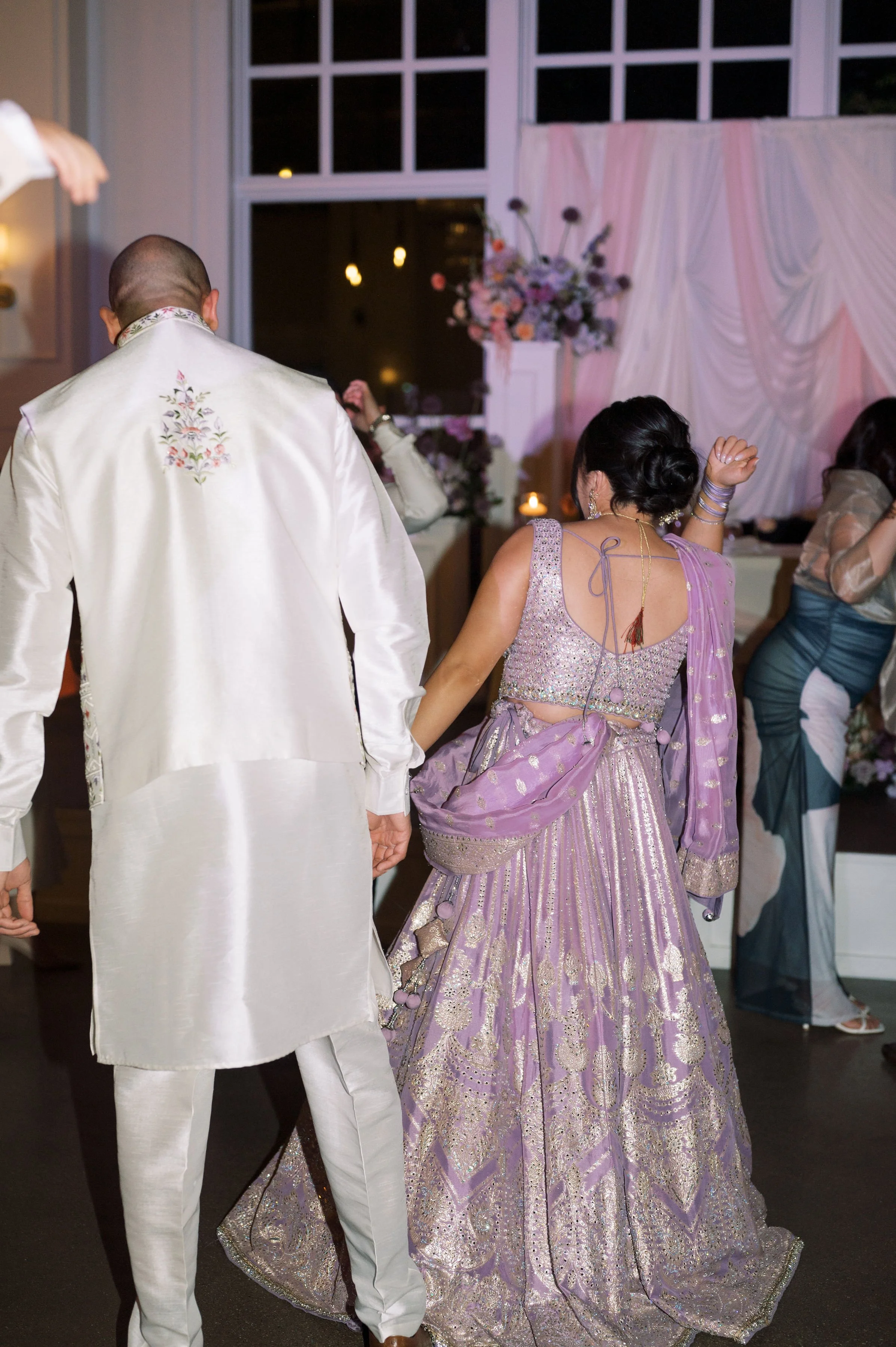 People dancing at a celebratory event, with women in traditional Indian attire and a man in white traditional clothing, at night with floral decorations.
