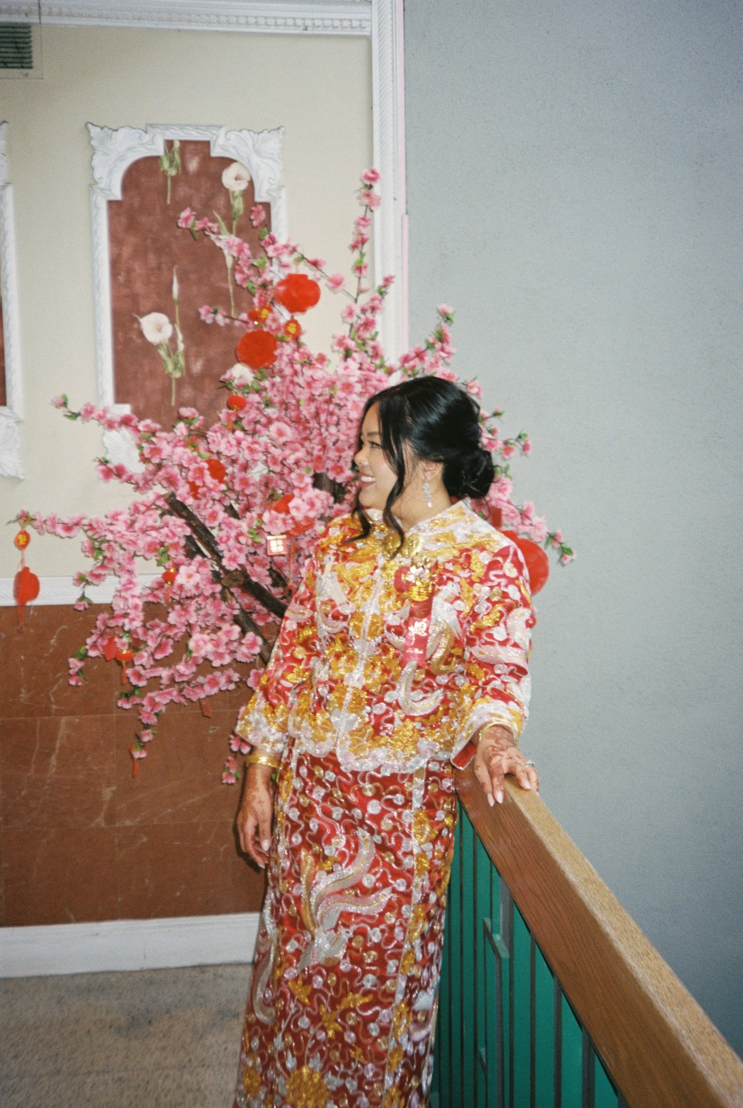 A woman in traditional red, gold, and white embroidered attire smiling, standing beside a pink cherry blossom tree with hanging red decorations, indoors with a light-colored wall and decorative wall art.