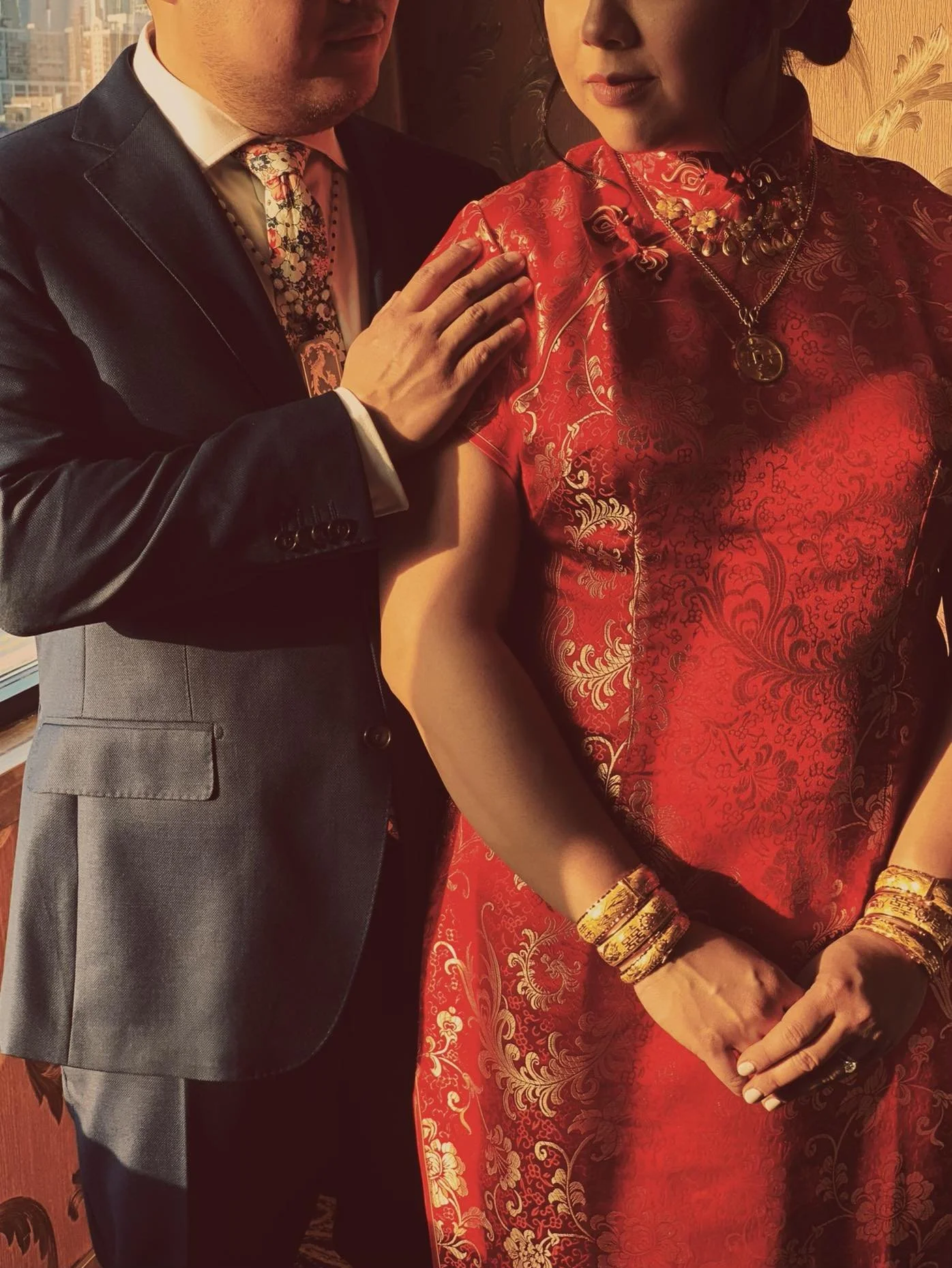 A man and a woman dressed in formal attire, with the man holding the woman's shoulder, is shown in this photo. The woman is wearing a traditional red silk dress with gold jewelry, and the man is wearing a suit with a colorful tie. The setting appears