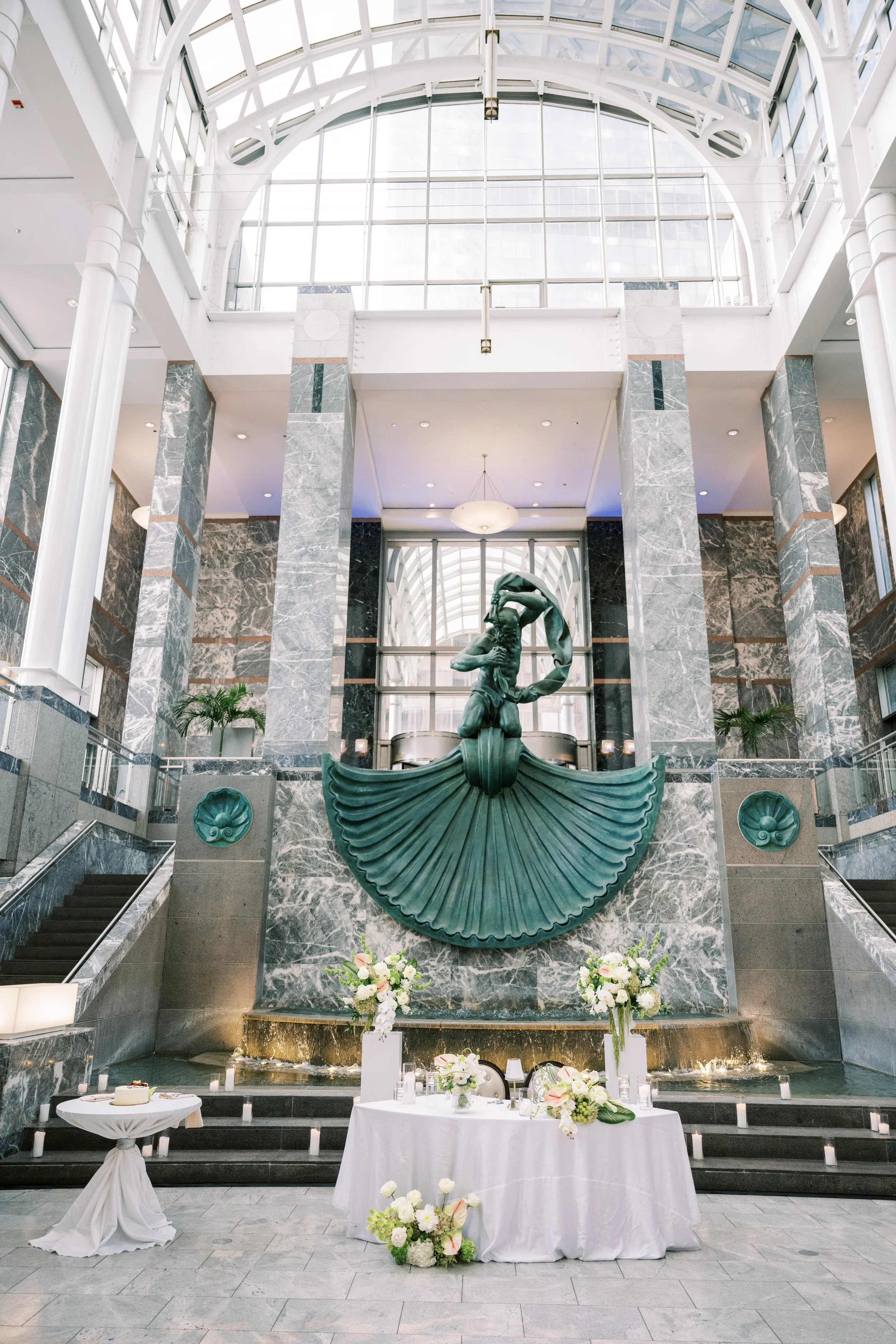 Elegant indoor space with a large arched glass ceiling, marble columns, and a central bronze sculpture of a woman. In front of the sculpture, there is a table with a white tablecloth, floral arrangements, and a birthday cake, indicating a celebration
