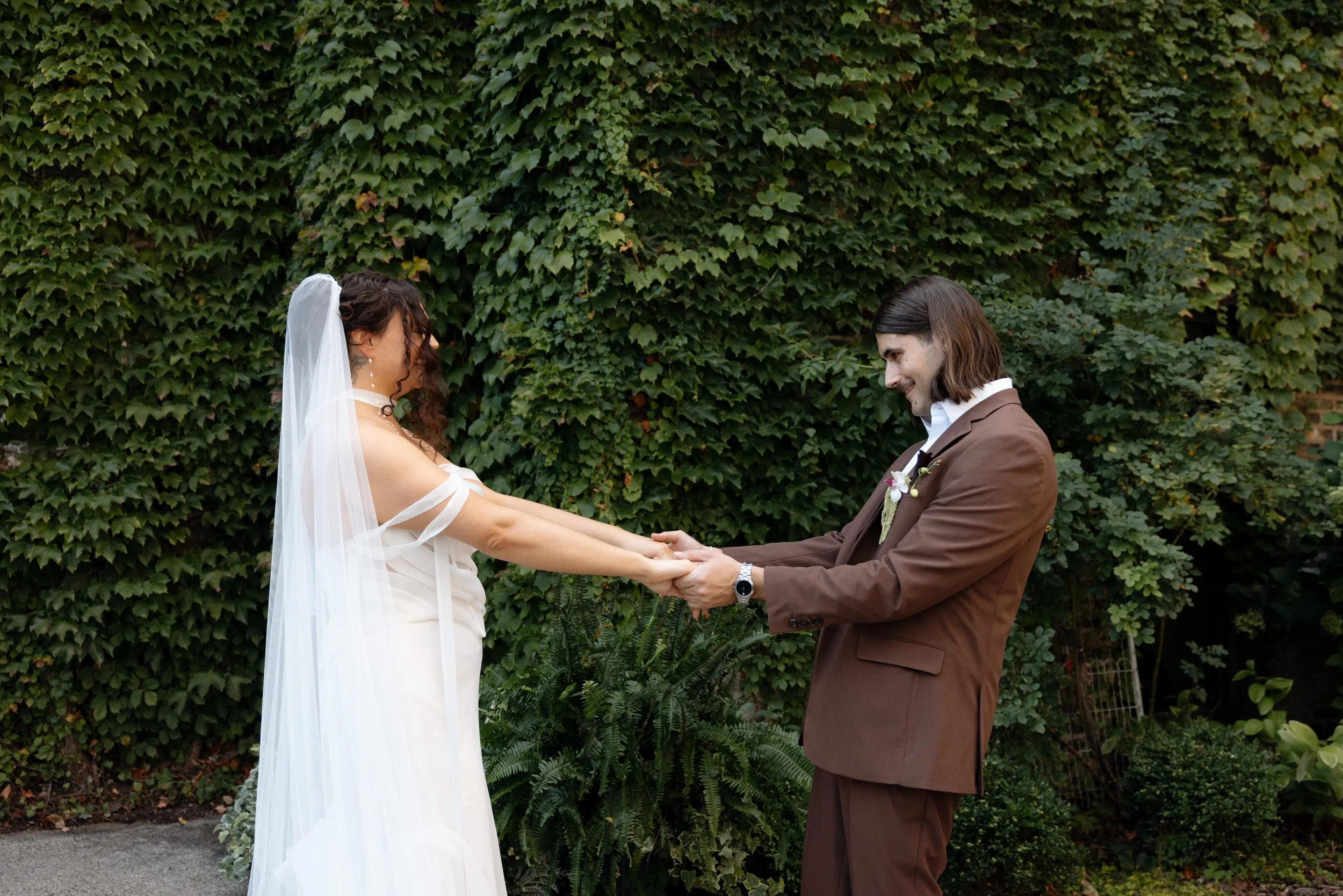 A bride and groom holding hands during their wedding ceremony outdoors in front of a green ivy-covered wall.