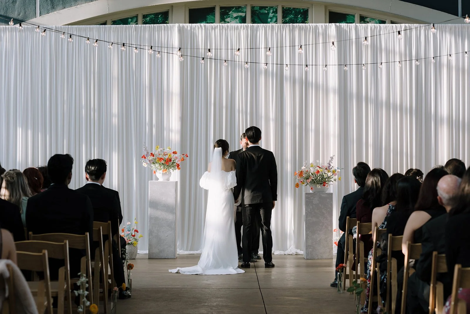 Bride and groom exchanging vows during a wedding ceremony in front of a white curtain backdrop with flowers