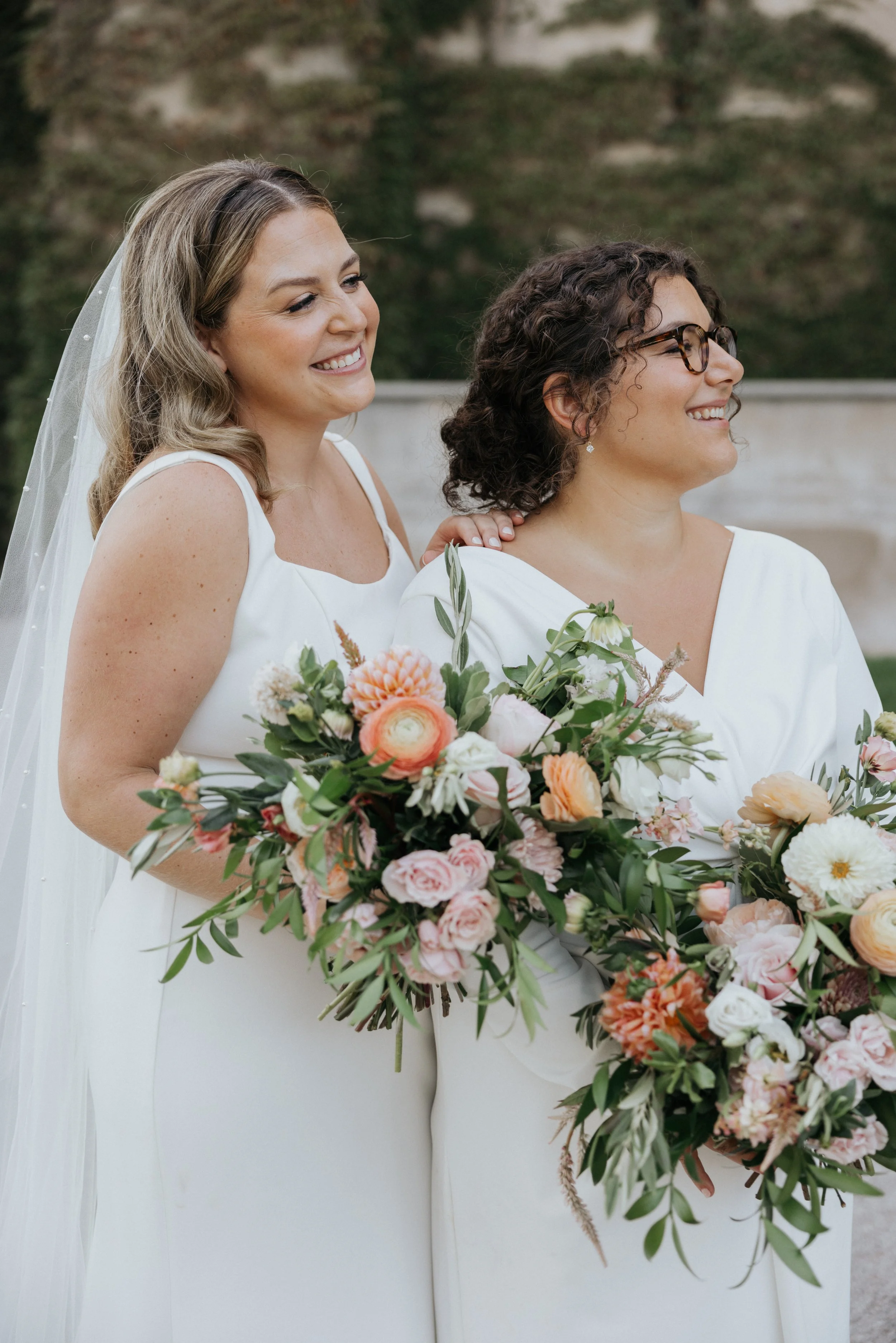 Two women in white dresses at a wedding, holding a large bouquet of pink, white, and peach flowers with greenery.