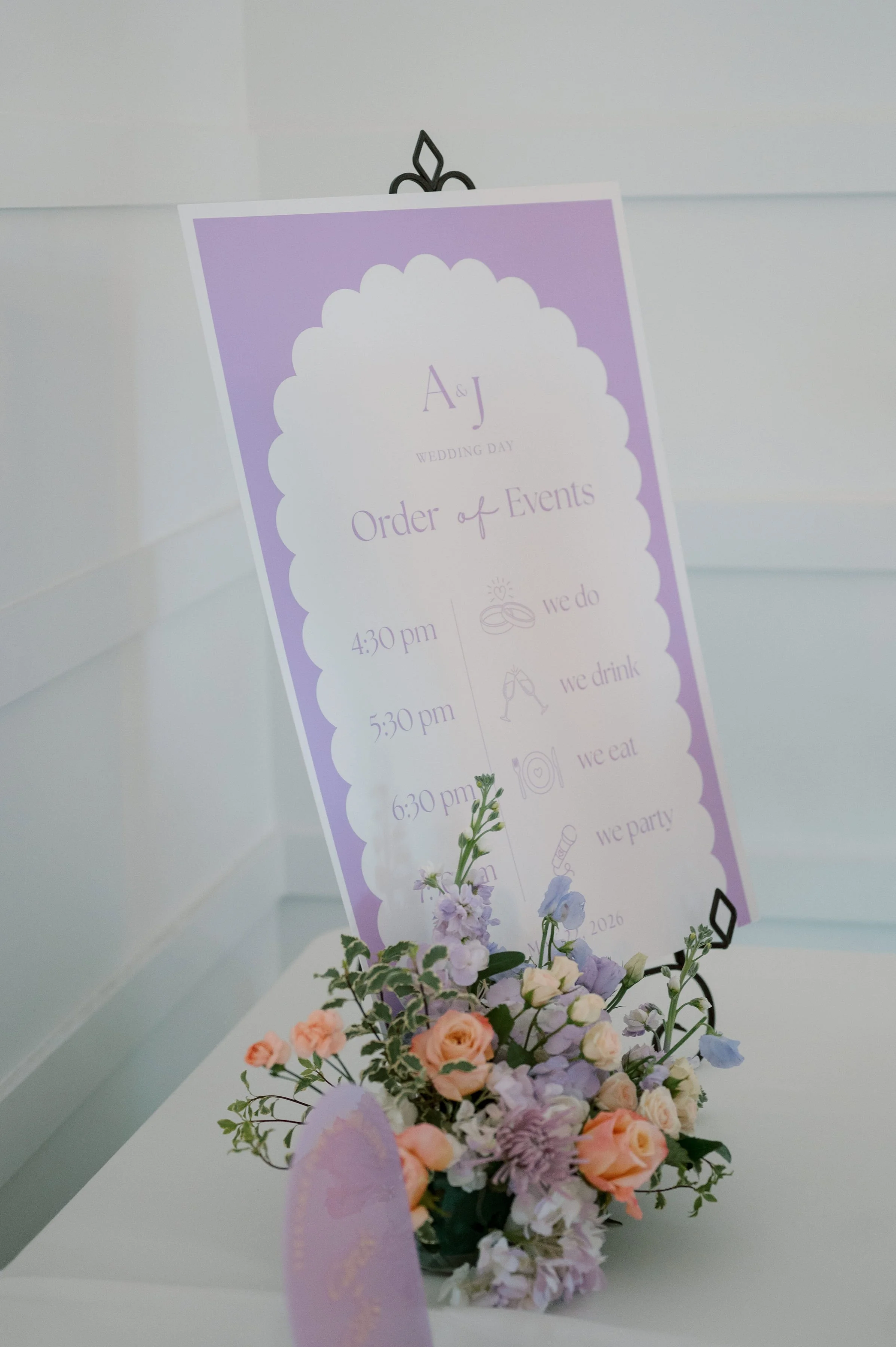 A wedding event schedule sign in purple and white, displayed with a flower arrangement at the bottom, featuring various flowers and greenery.