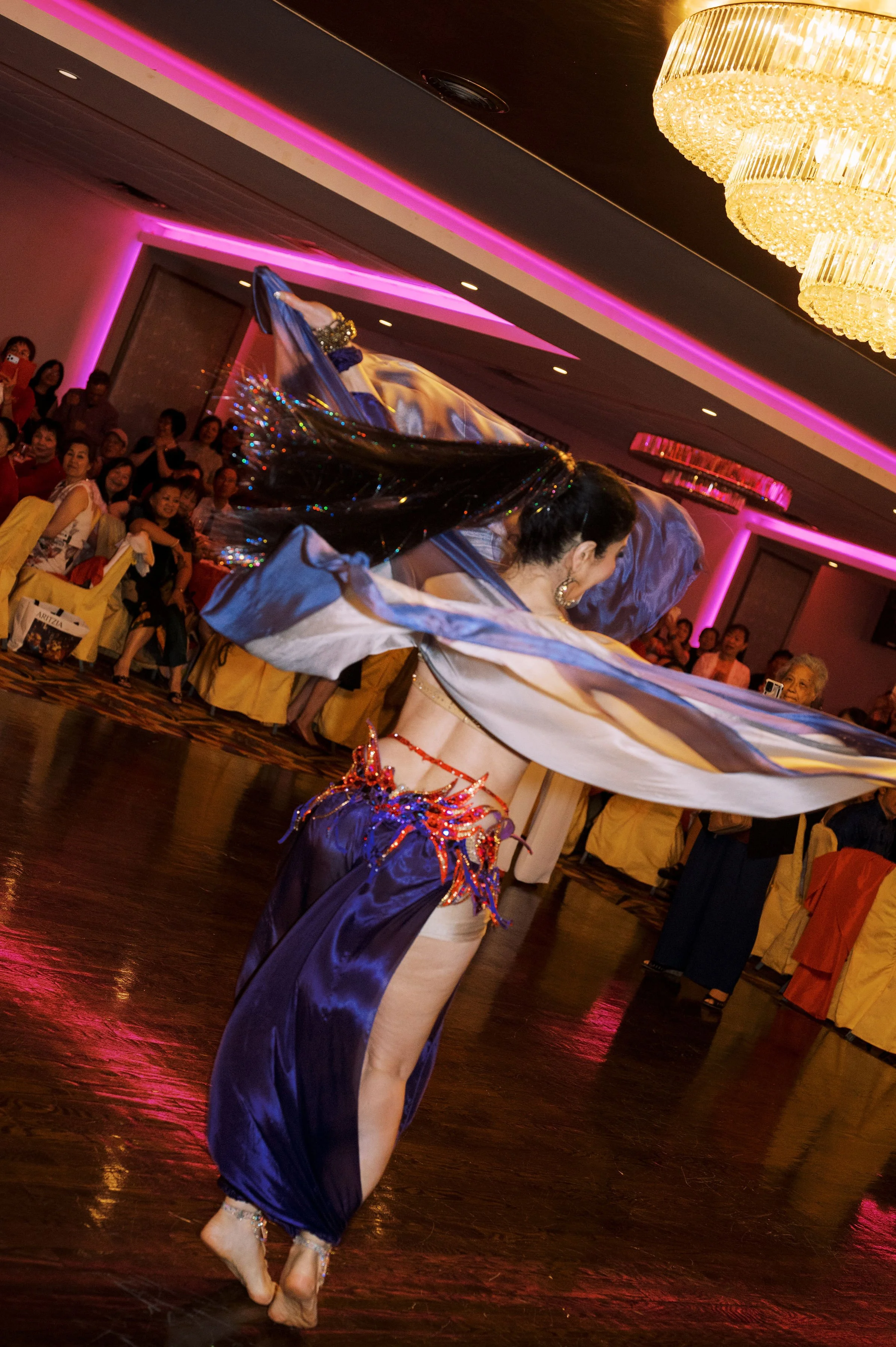 A dancer in a purple costume performs a dance with a flowing fabric in a banquet hall, with an audience watching and taking photos. The hall has purple lighting and large chandeliers.