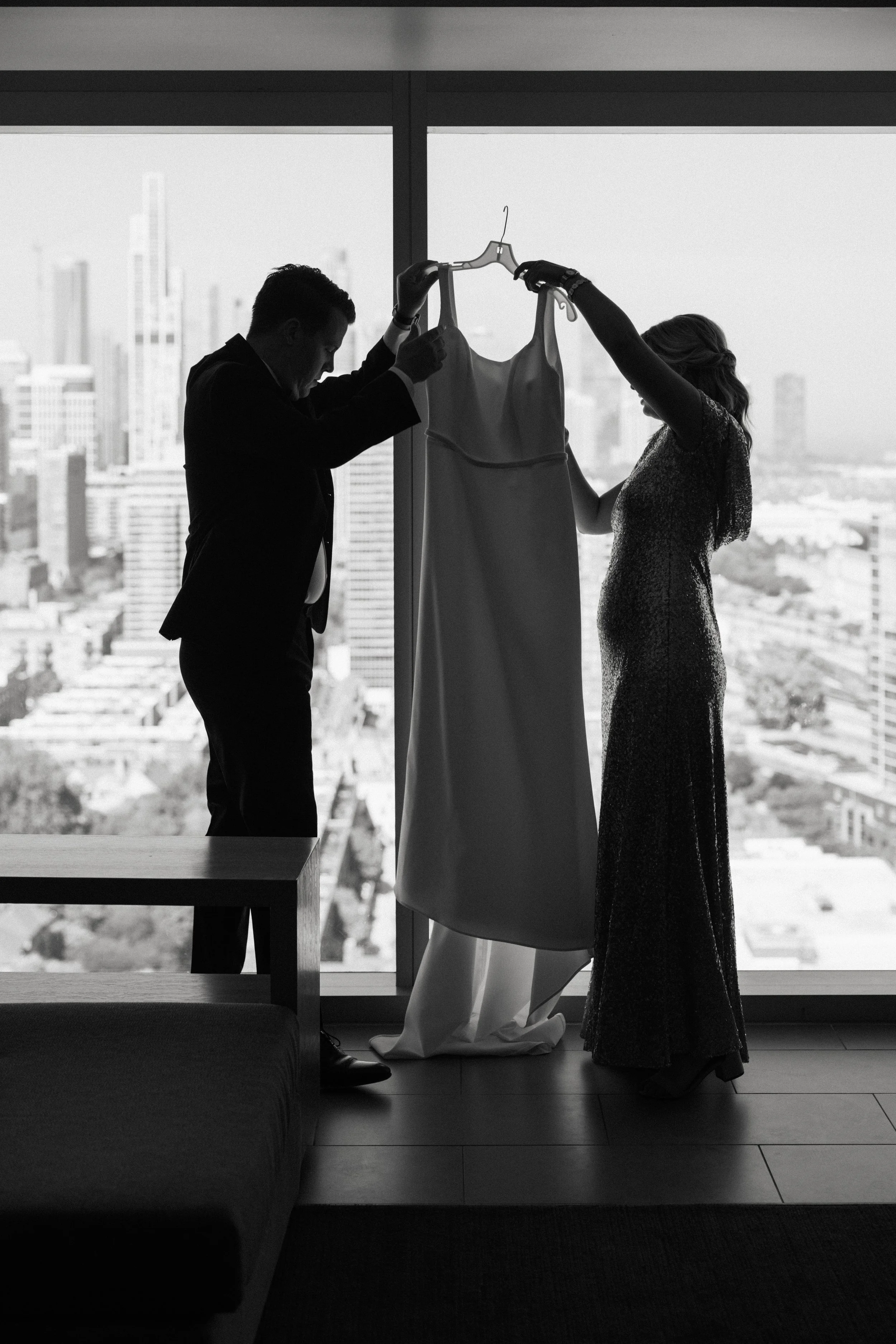 Black and white photo of a man and a woman in formal attire holding up a wedding dress on a hanger inside a high-rise building with a city skyline in the background.