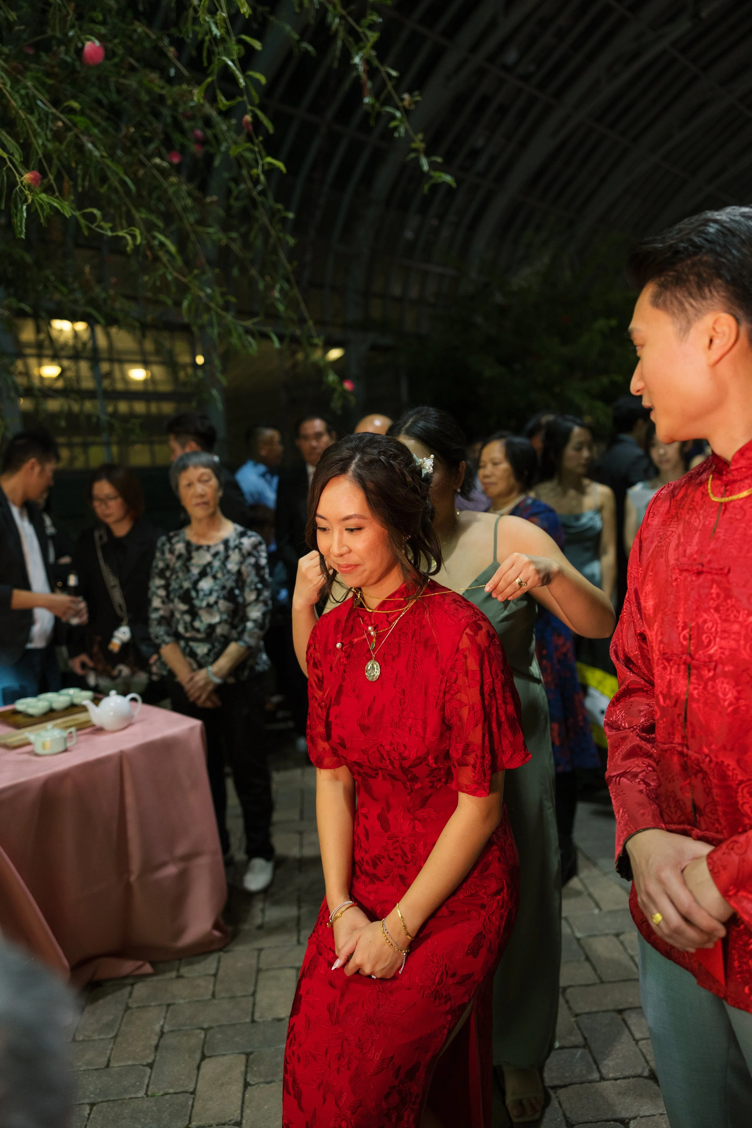 A group of people at a social gathering, with a woman in a red dress and jewelry in the foreground, and other people in the background, some wearing traditional Asian attire.