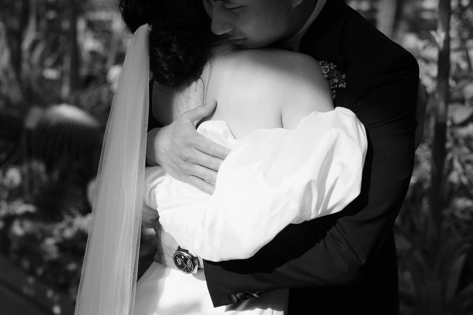 A black-and-white photo of a bride and groom hugging closely, with the groom embracing the bride's shoulder and back, wearing a white dress and veil, outdoors in a natural setting.
