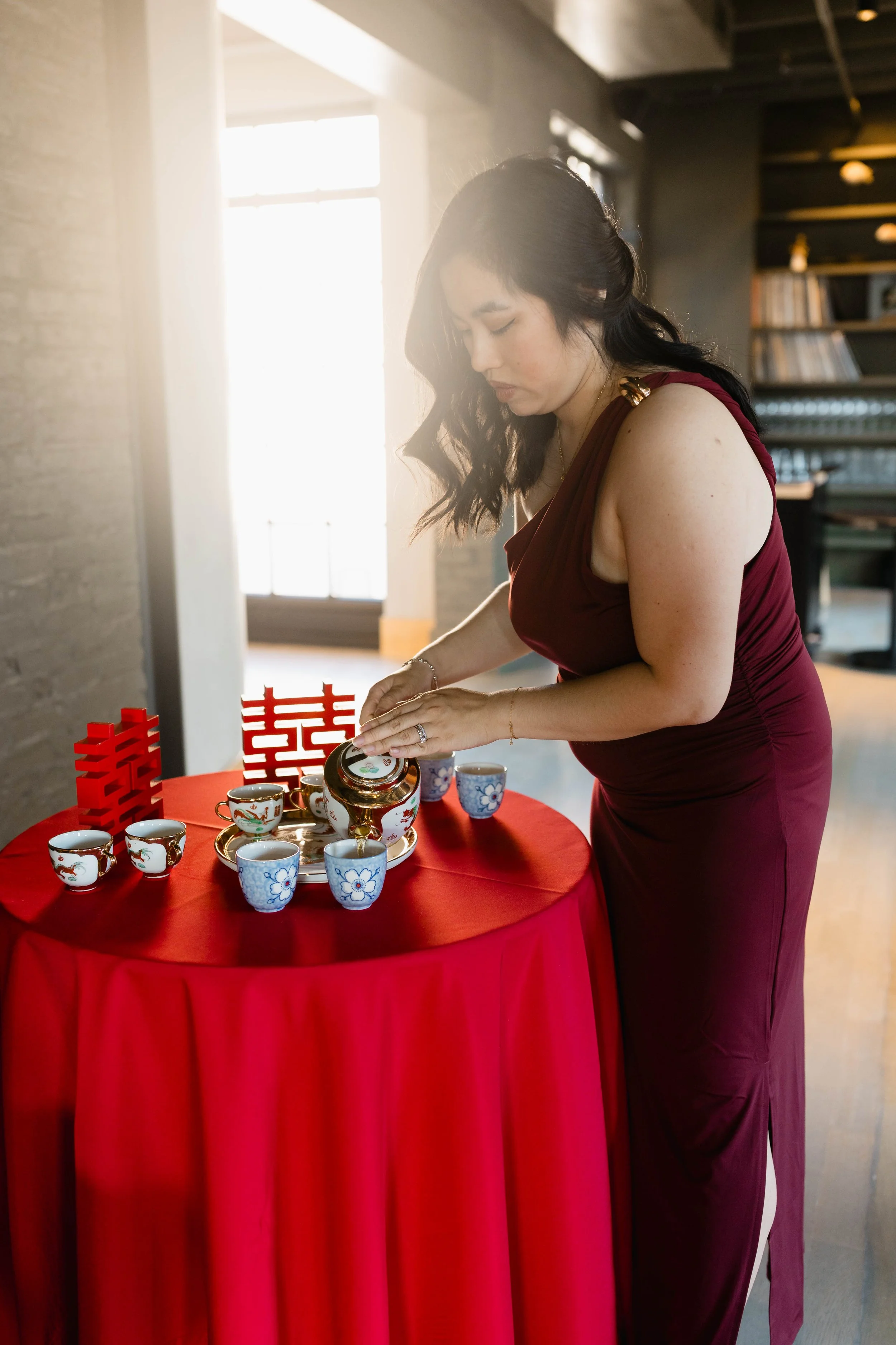 A woman in a deep red dress is pouring tea into teacups on a red-draped round table, displaying Chinese traditional decorations and cups, inside a modern room with large windows and bookshelves.