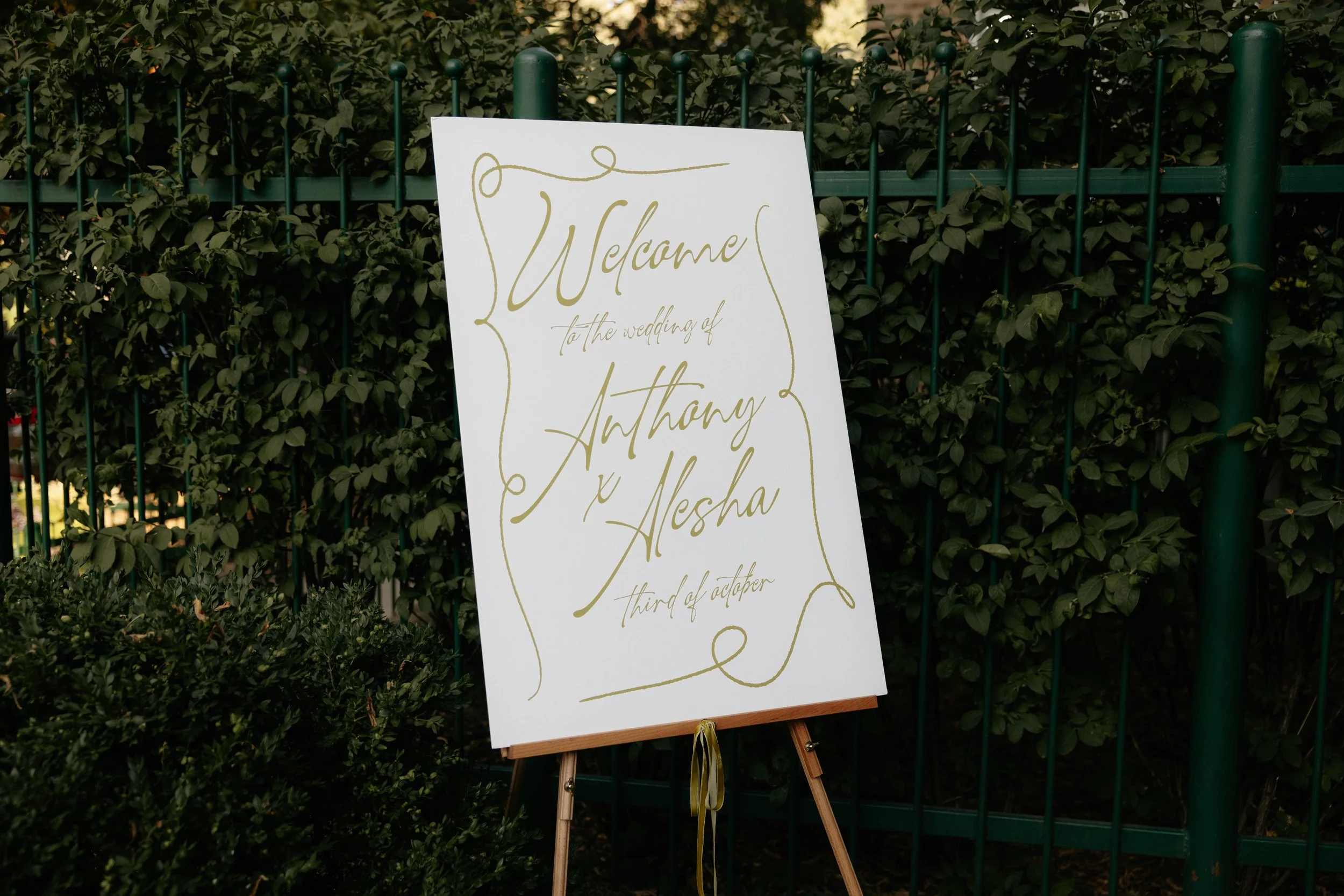 A wedding welcome sign on an easel, with decorative gold writing, placed in front of a green hedge and a green metal fence.