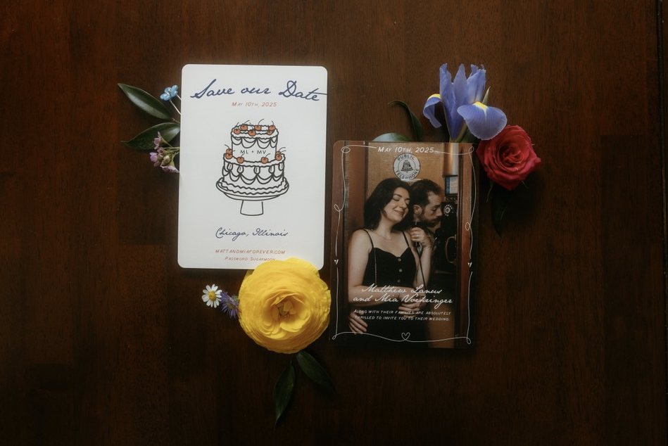 Wedding save-the-date card, wedding invitation with a photo of a woman and man, and arranged flowers including purple irises, a red rose, a yellow ranunculus, and small purple and white flowers, all on a wooden surface.