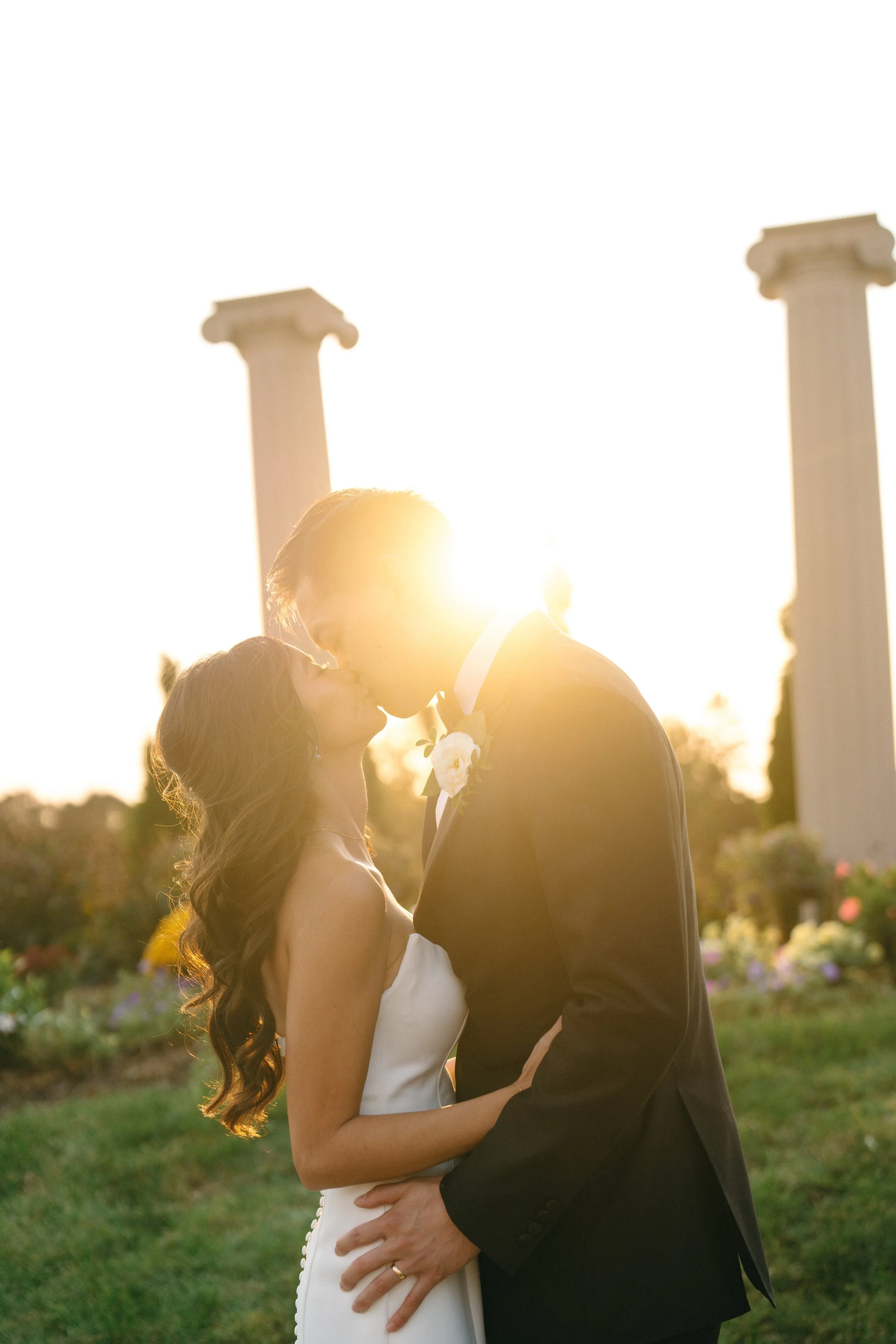 A bride and groom kissing outdoors during sunset, with ancient columns in the background.