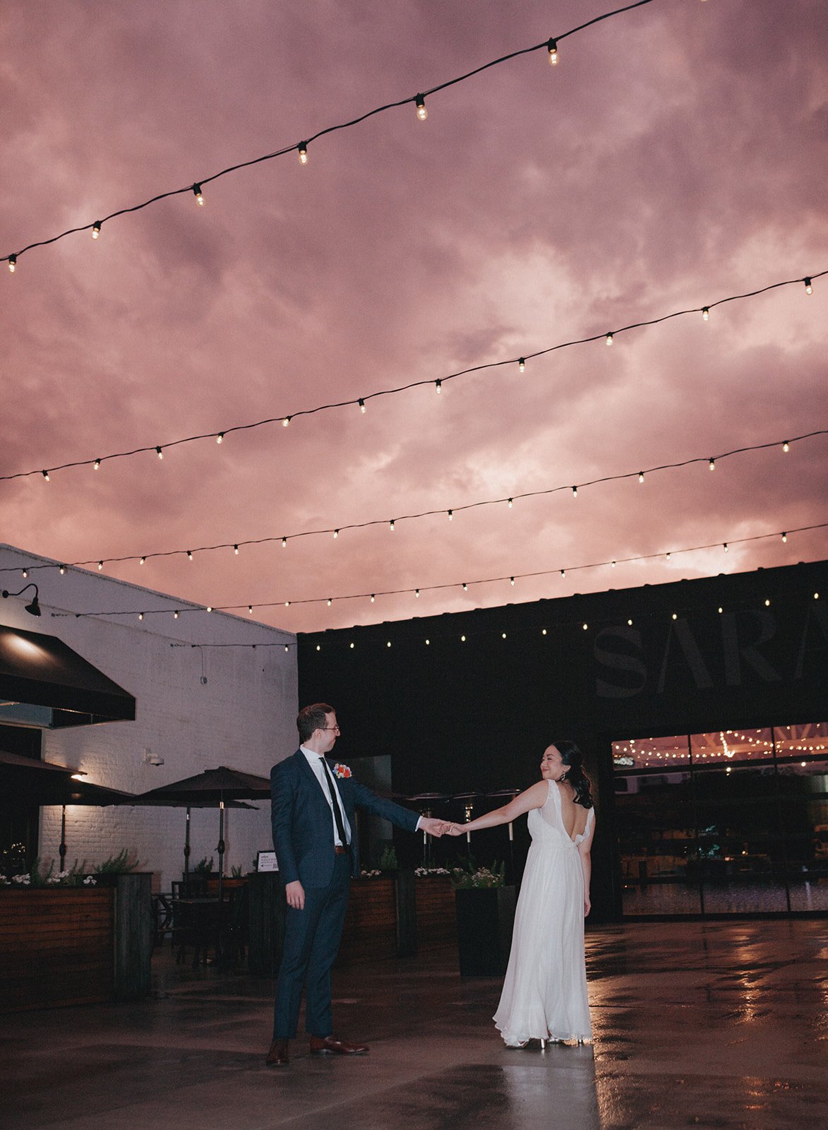 A couple dancing outdoors at sunset, holding hands and smiling. The sky is pink and purple with string lights hanging above them.