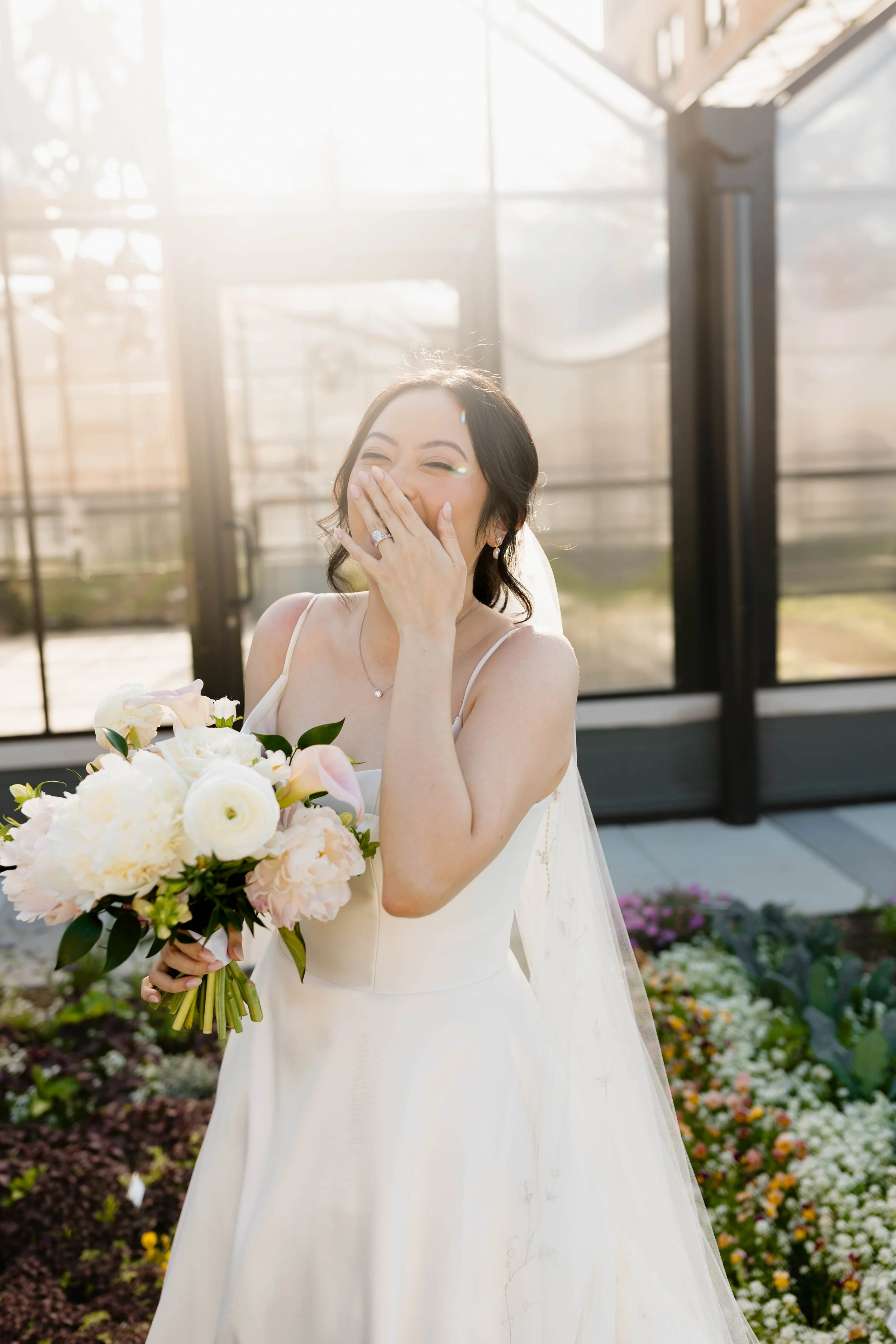 A woman in a white wedding dress holding a bouquet of white and blush pink flowers, covering her mouth in surprise and joy, standing in a brightly lit greenhouse with glass walls and a garden of colorful flowers in the background.