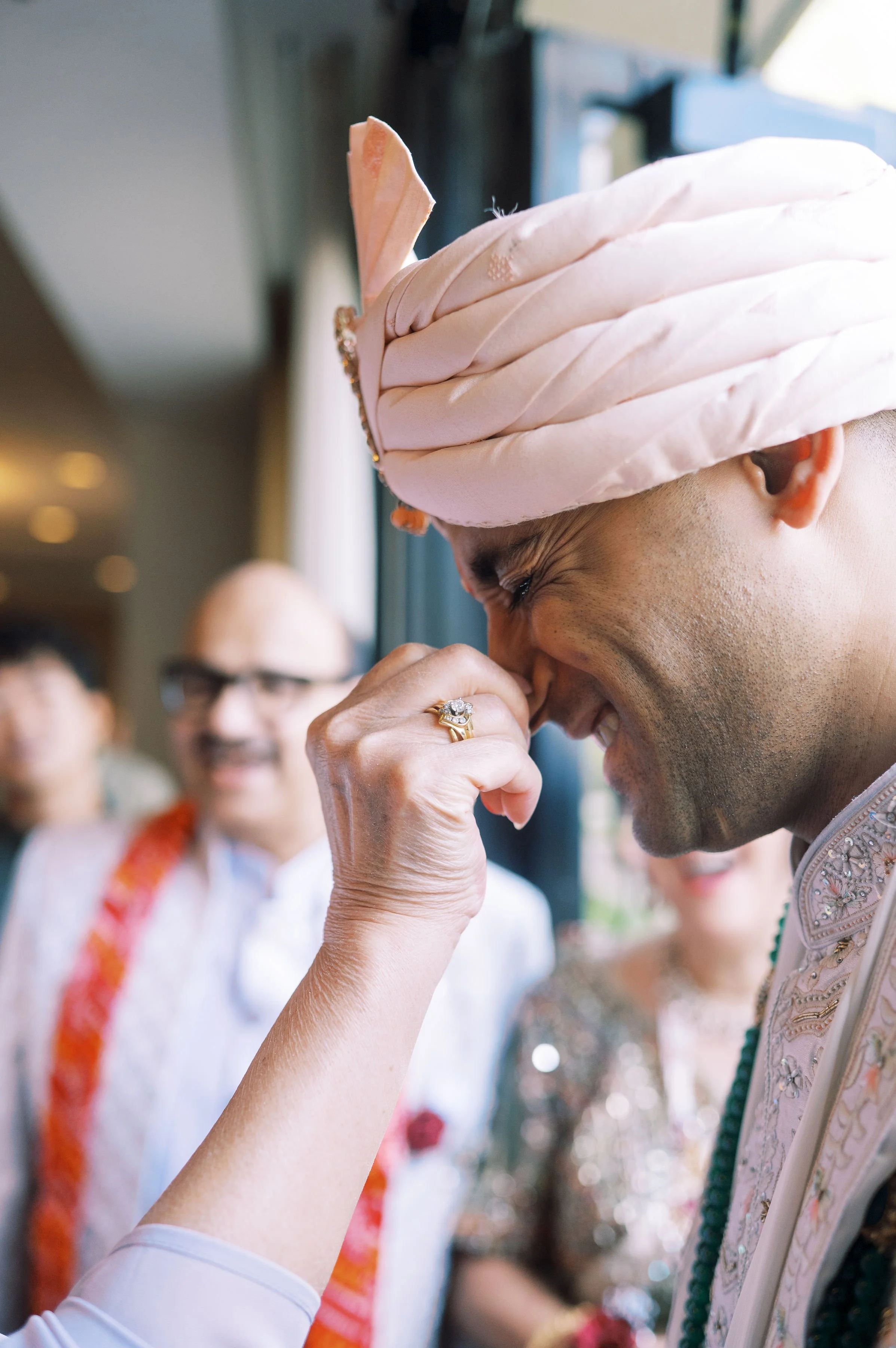 A man wearing a pink turban is sniffing a floral garland while a woman in white with glasses and a colorful sash is in the background, smiling. The scene appears to be a celebratory or ceremonial event.