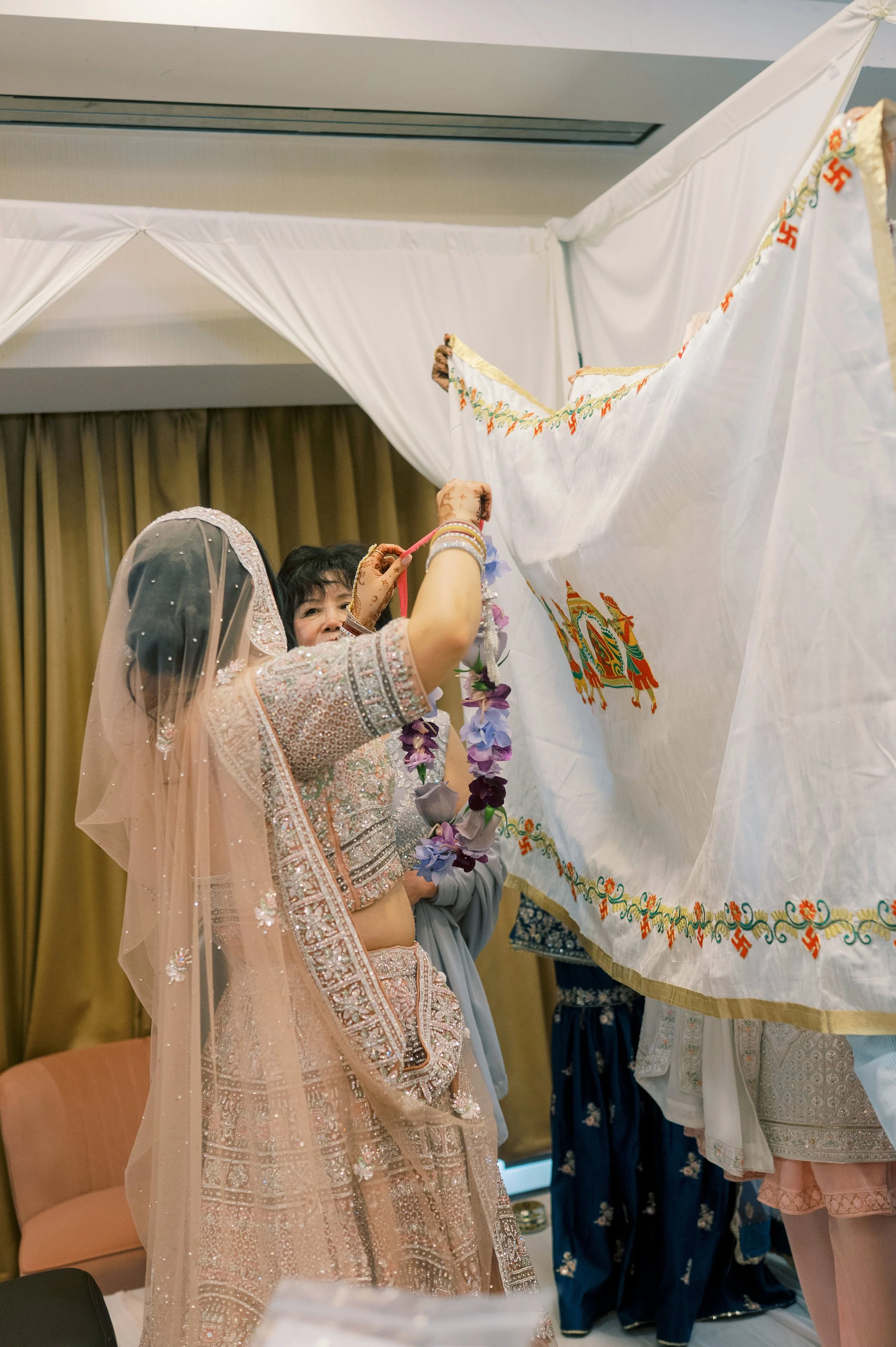Women dressed in traditional Indian attire, with henna on their hands, participating in a cultural ceremony involving a decorated white fabric with intricate designs and symbols, in an indoor setting.