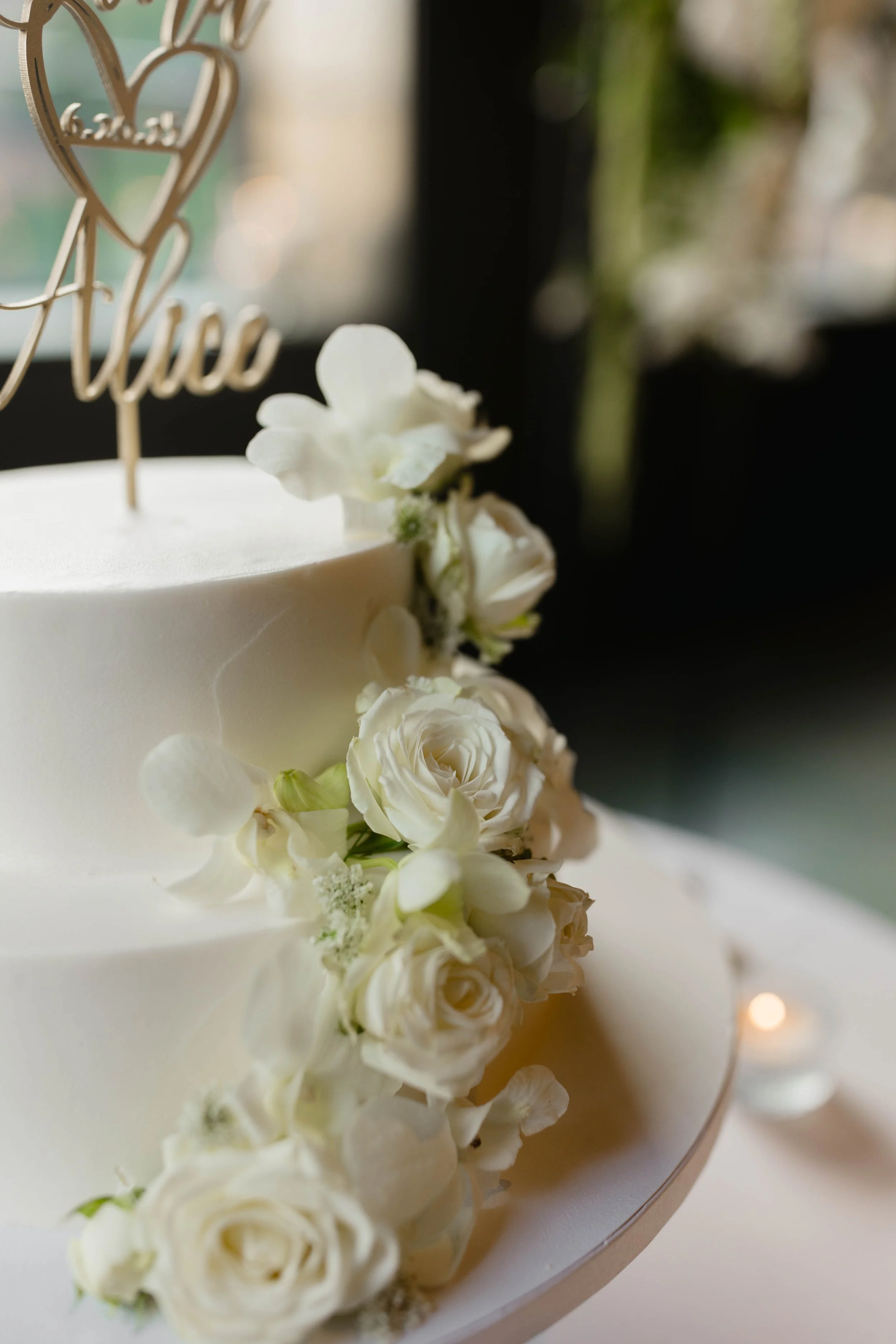 A white wedding cake decorated with white roses and small white flowers, with a gold topper that says 'love' in cursive.