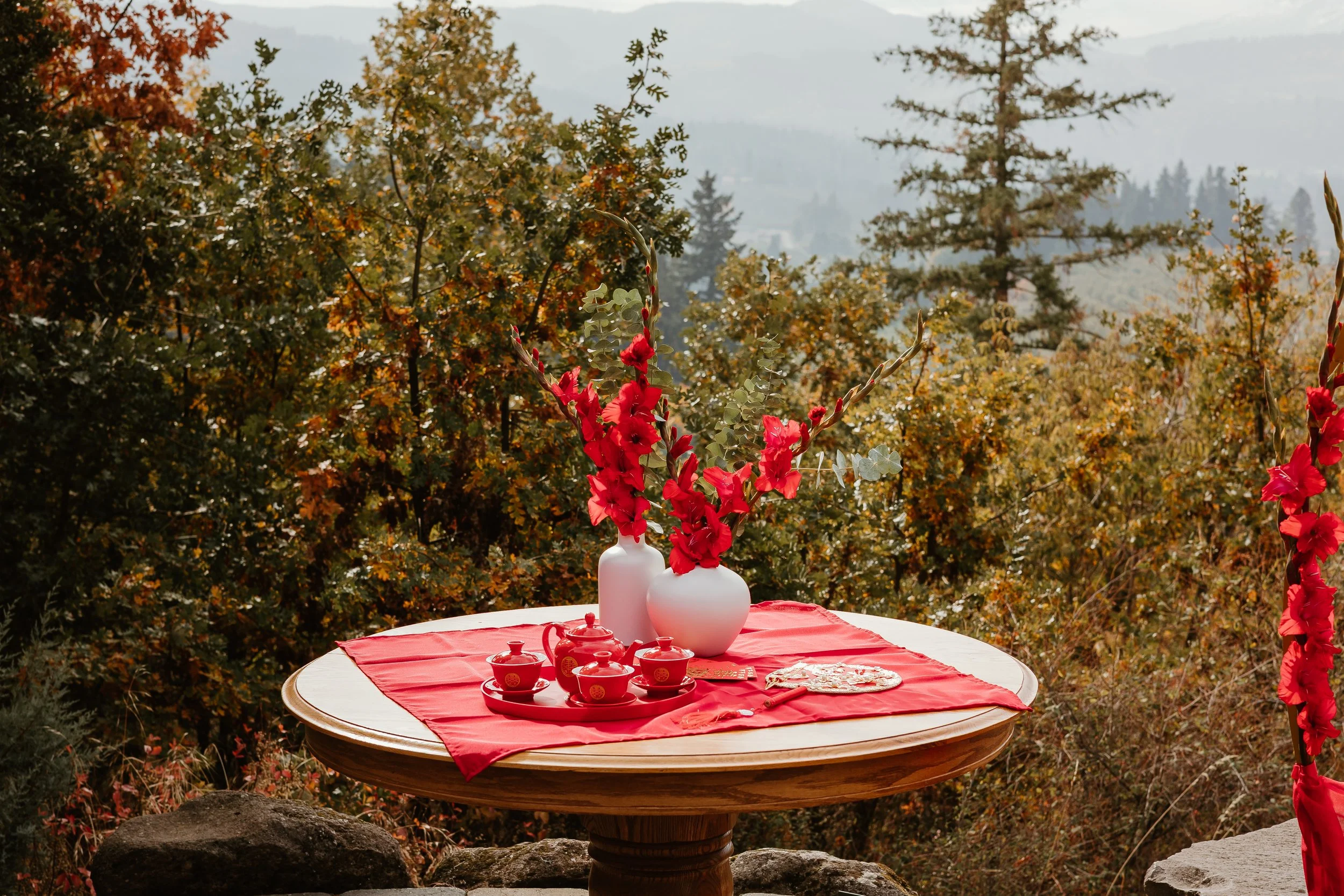 A round wooden table with a red tablecloth, decorated with white vases and red flowers, set for a tea or celebration outdoors, with a background of autumn foliage on trees.
