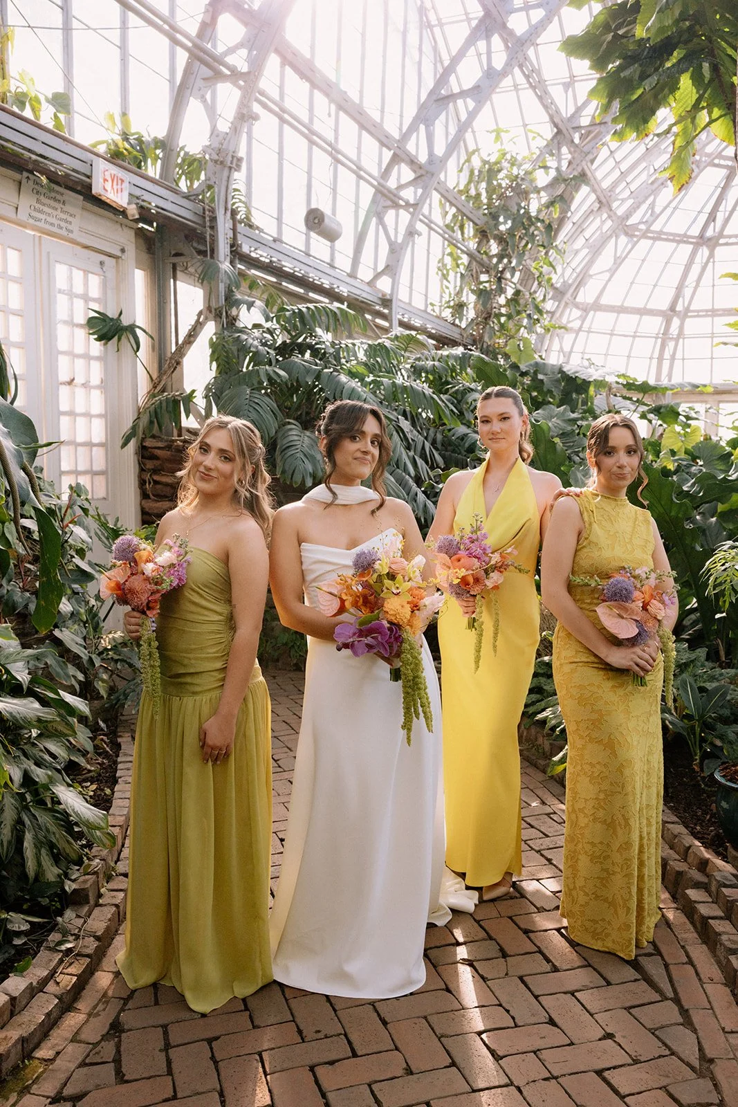 Four women in colorful dresses holding bouquets of flowers, standing in a greenhouse filled with lush green plants and bright sunlight.