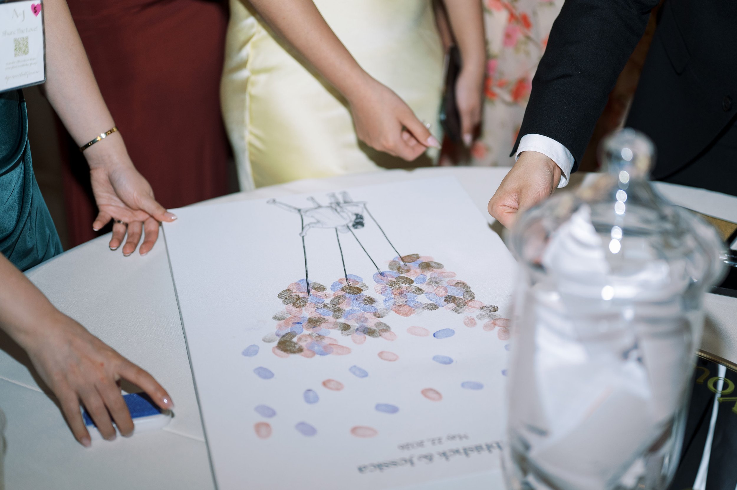 Close-up of a group of people gathered around a table, examining a large sheet with a drawing of a person and colorful dots. One individual is pointing at the sheet, and a water bottle is partially visible on the right side.