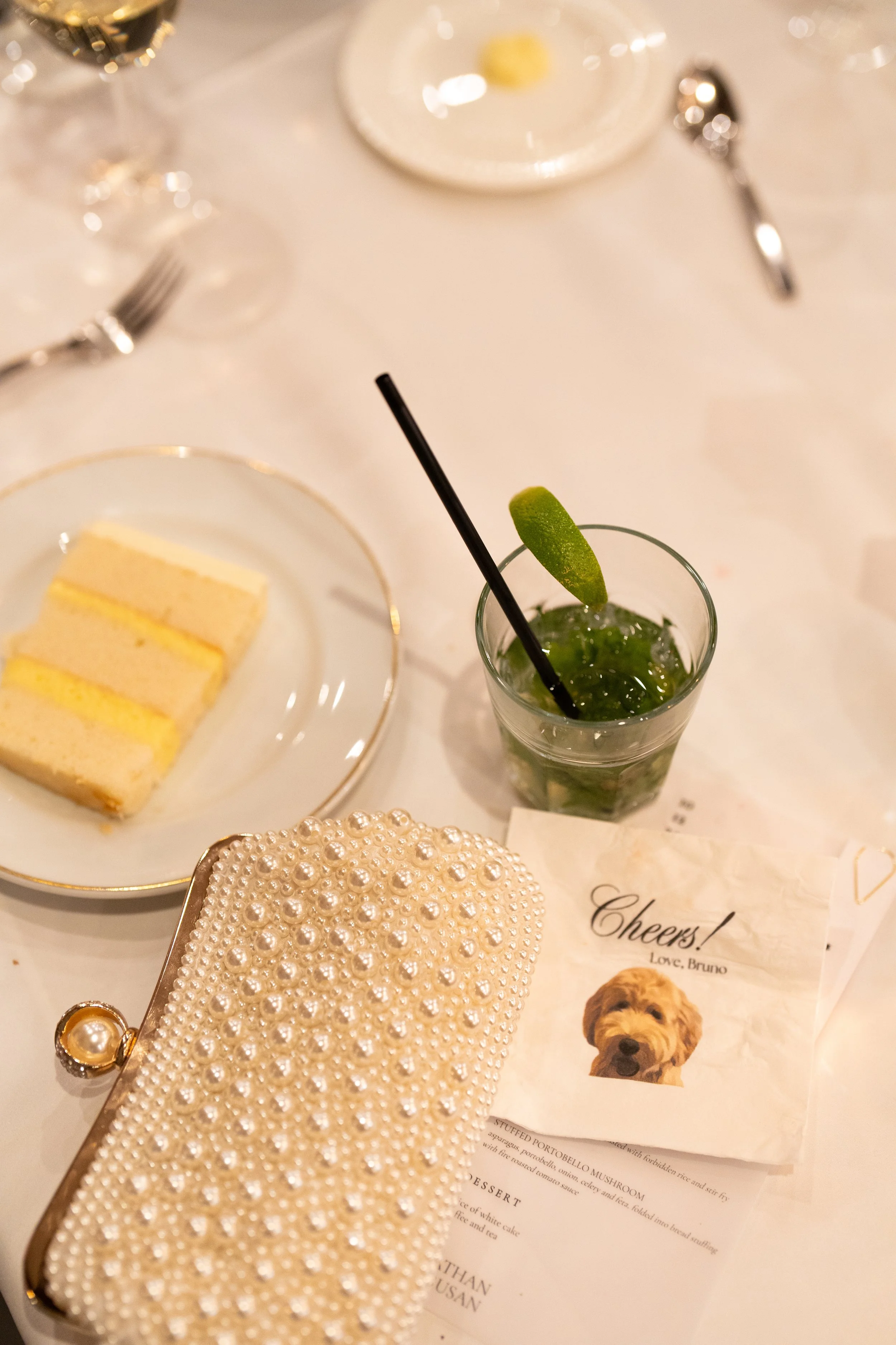 A table setting with a glass of mint mojito, a plate with slices of cake, a pearl-studded clutch purse, and a dog-themed greeting card.