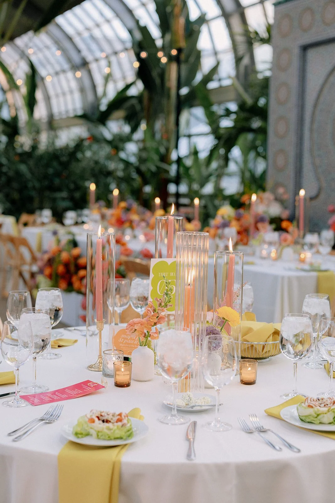 Elegant banquet table set with pink candles, flowers, glassware, and yellow napkins in a greenhouse-like setting with lush greenery and string lights.