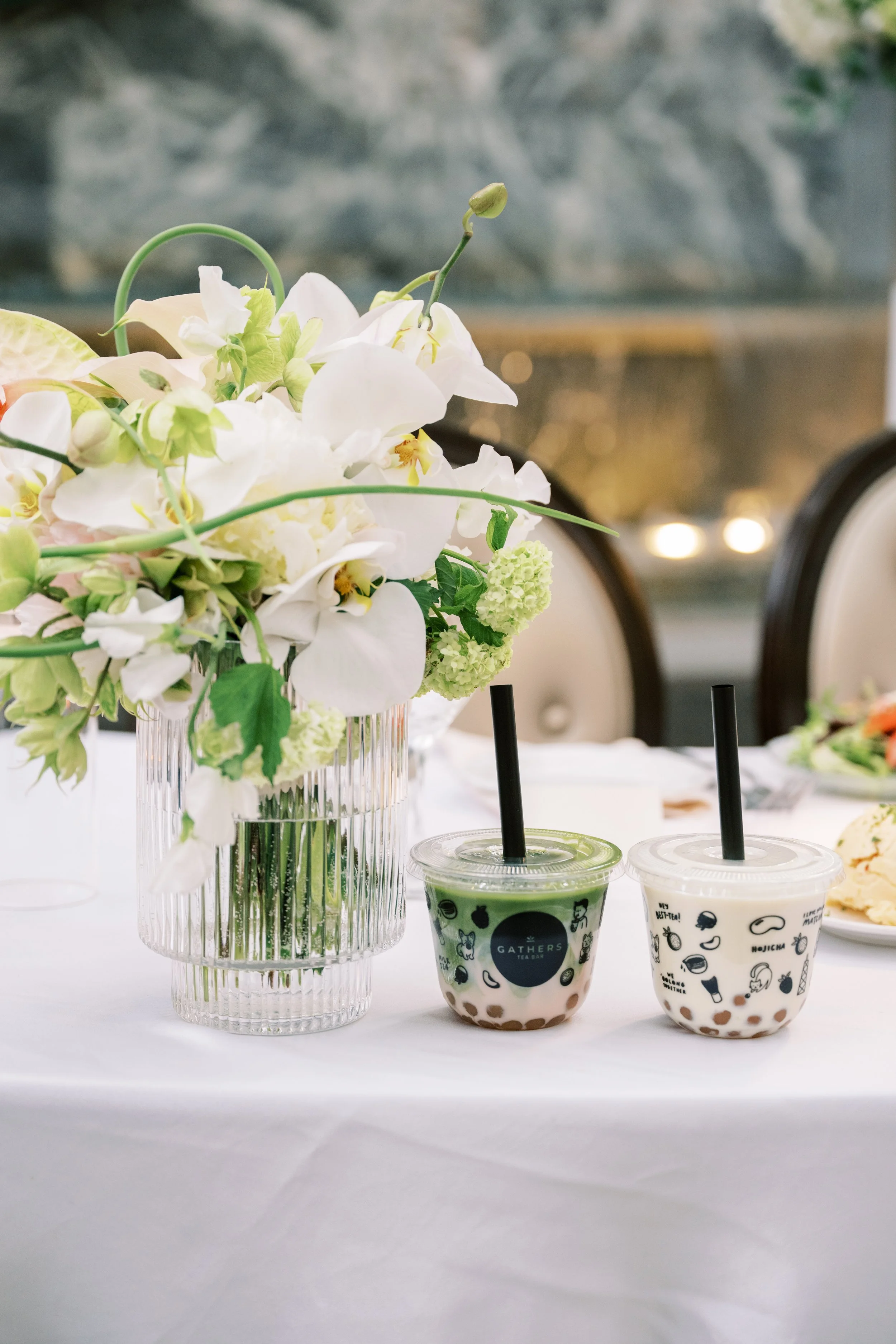 A floral centerpiece with white flowers and green accents in a glass vase, with two bubble tea drinks, one green and one white, on a white tablecloth in an indoor setting.