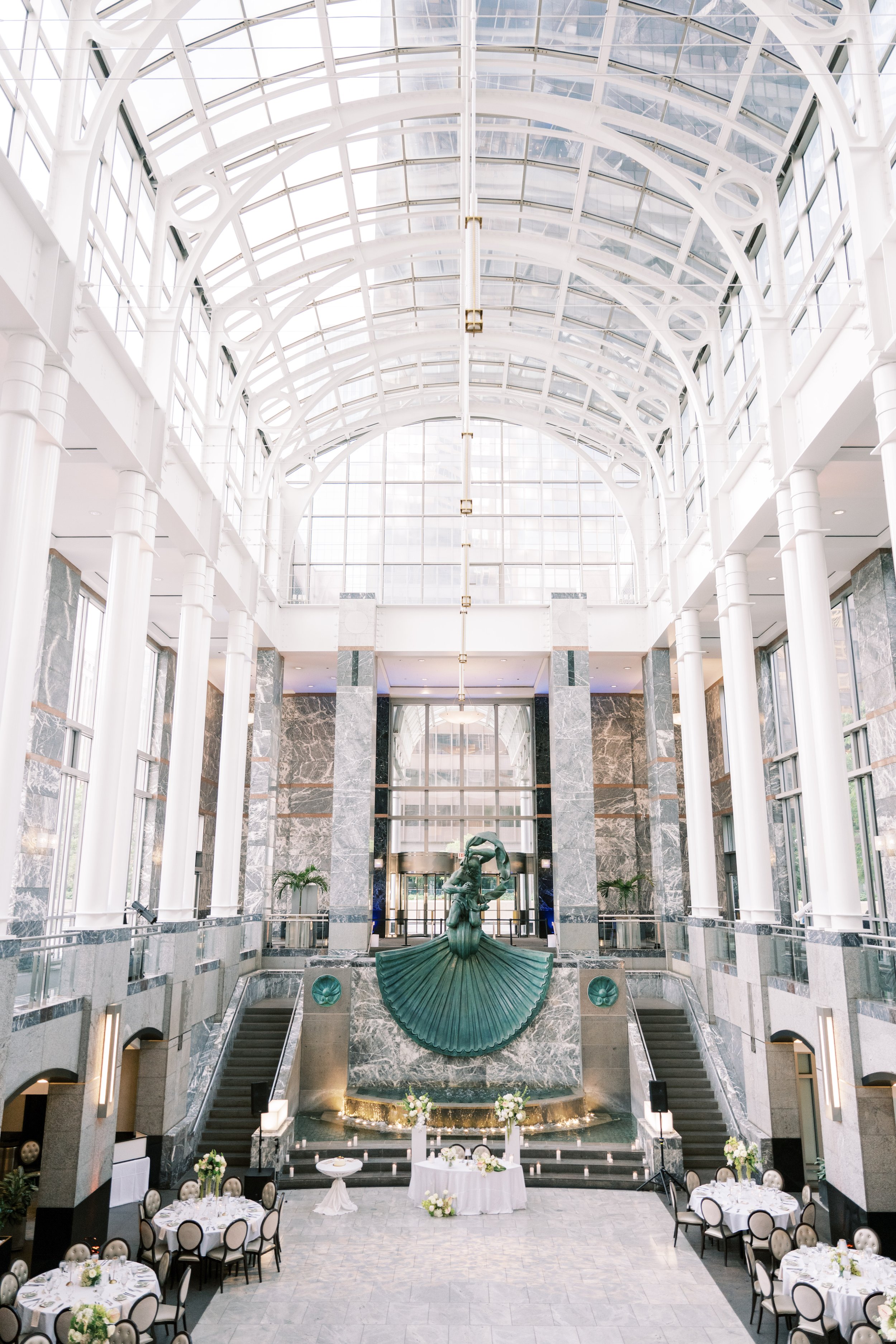 A spacious indoor atrium with a glass ceiling and white metal arches. In the center, there is a fountain with a sculpture of a woman holding a large shell-shaped object. Surrounding the fountain are decorated tables set for an event, with white table