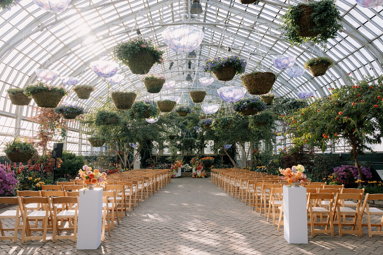 A greenhouse decorated for a wedding or event with rows of wooden chairs, floral arrangements, hanging flower baskets, and illuminated umbrella-shaped lights.