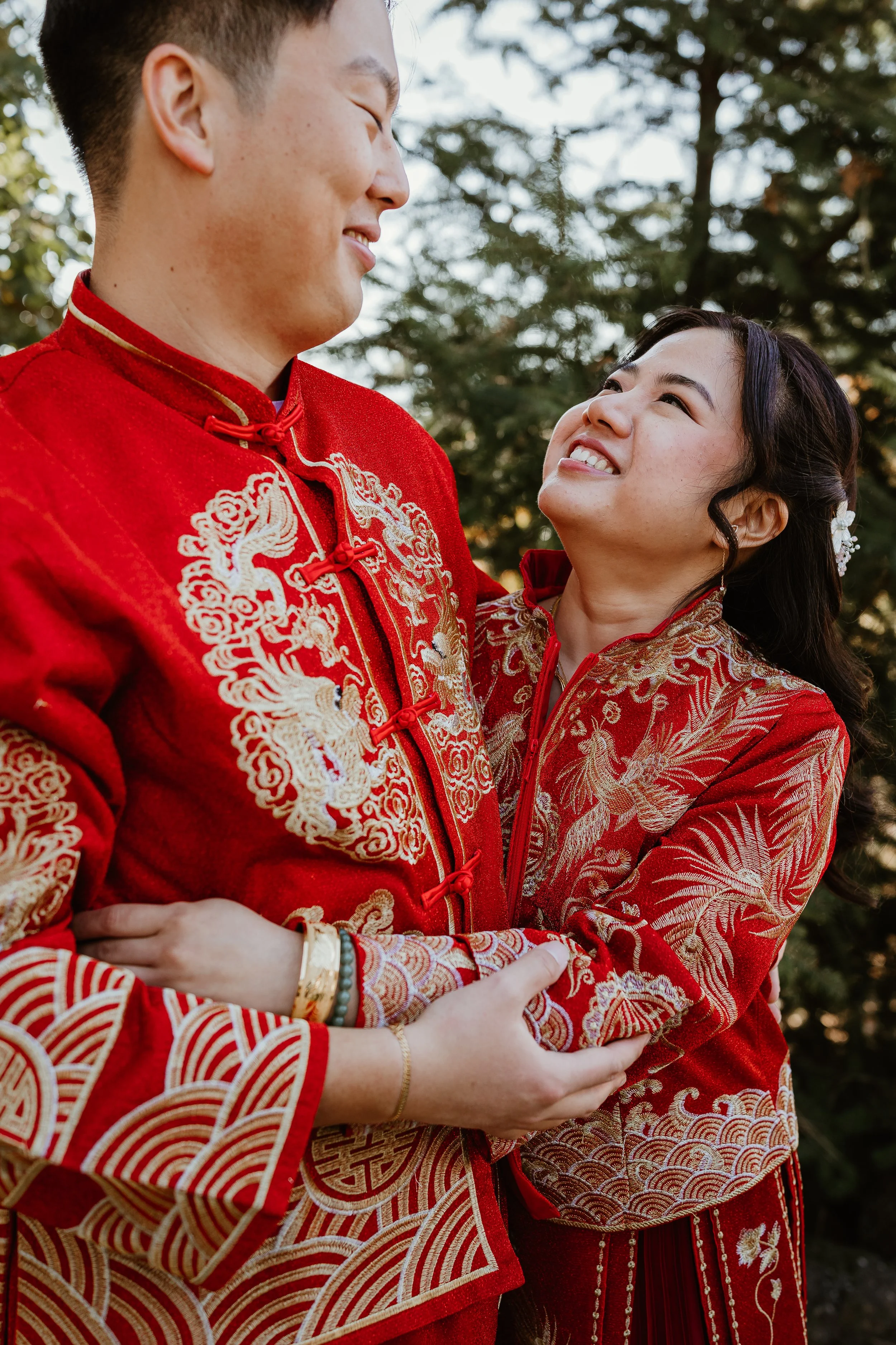 A couple wearing traditional Chinese wedding attire, embracing outdoors with trees in the background.