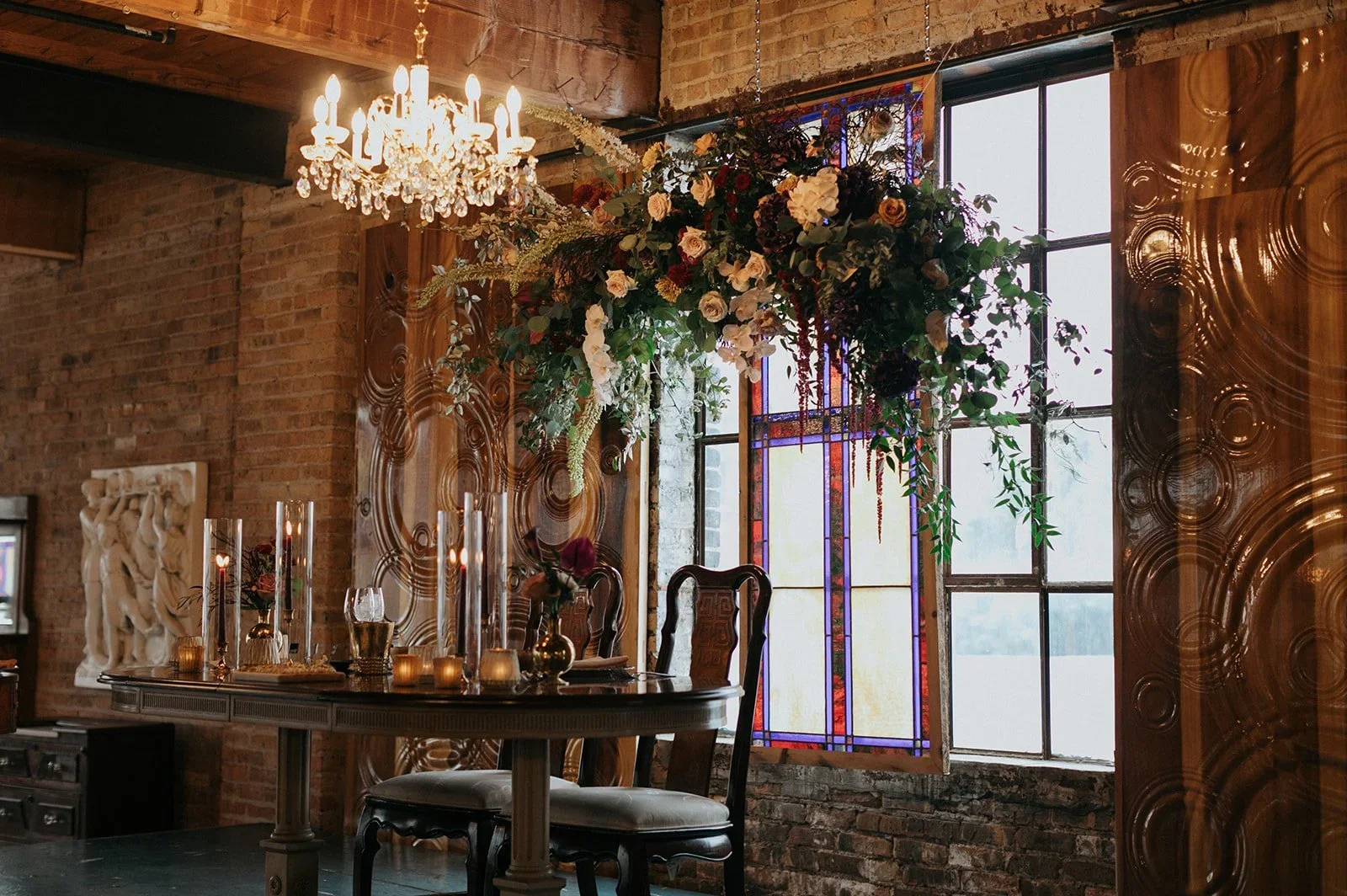 Interior of a brick wall room decorated for a special occasion with a chandelier, a floral arrangement hanging from the ceiling, a stained glass window, and a dining table with candles and flower arrangements.