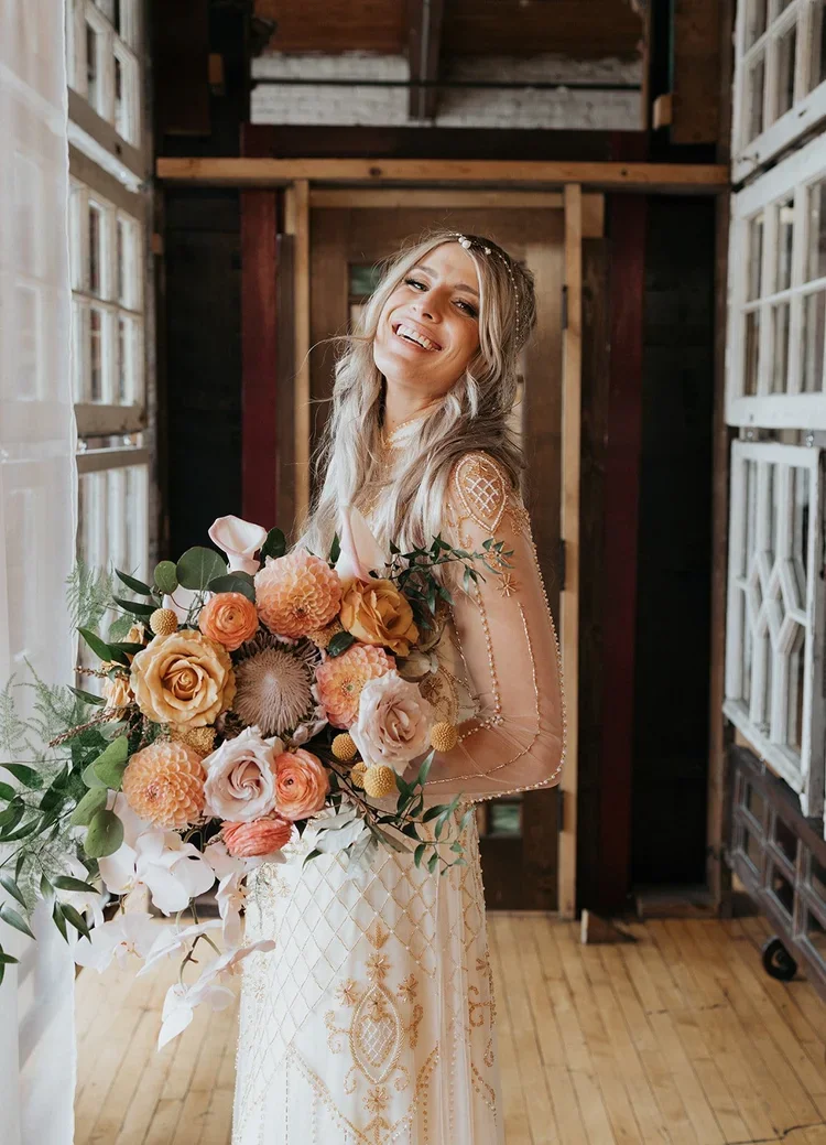 A smiling woman holding a large bouquet of peach, white, and pink flowers, standing indoors in front of wooden walls and white window frames, wearing a beige dress with intricate gold embroidery.