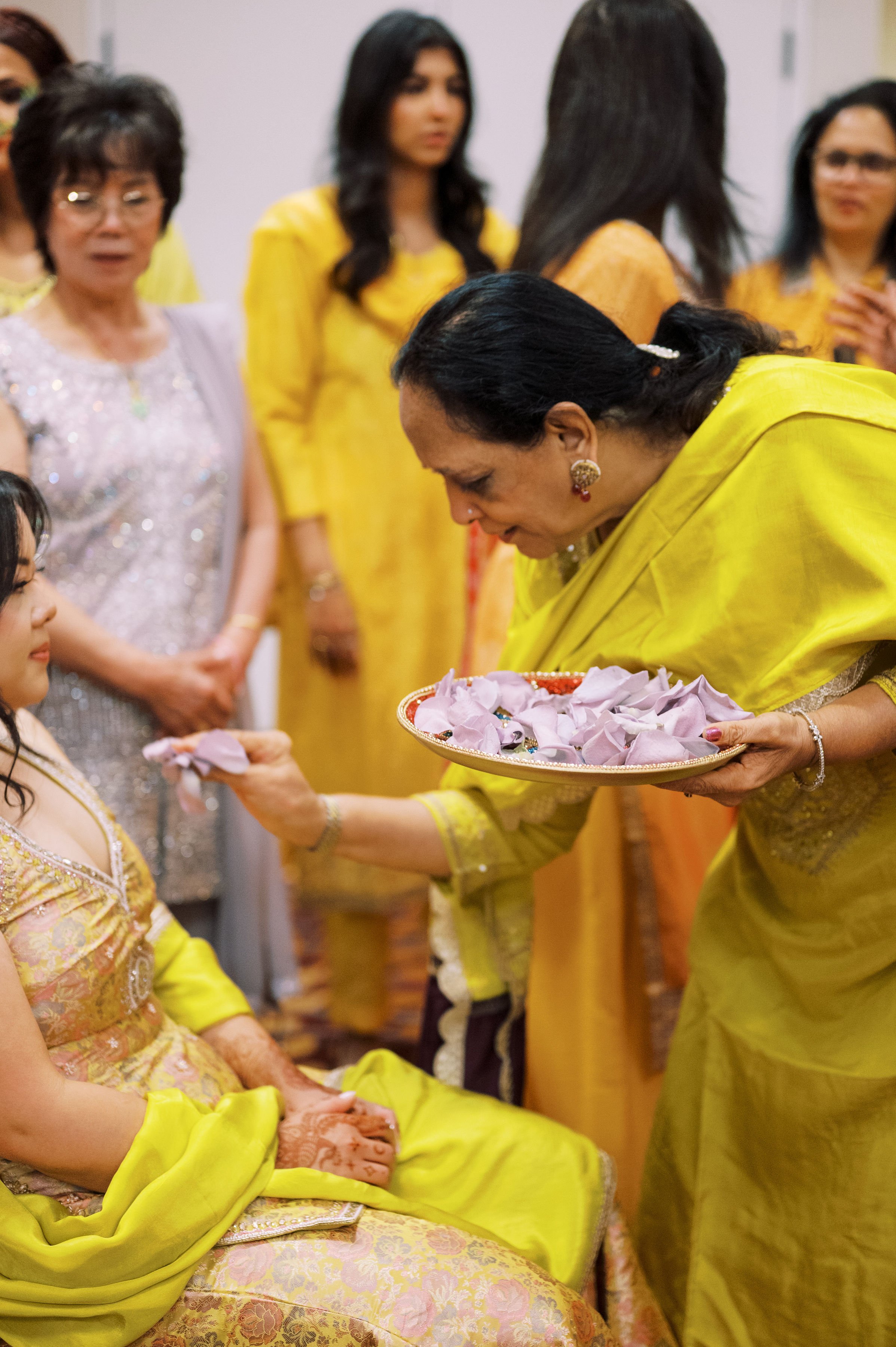 Group of women participating in a traditional Indian ceremony, dressed in colorful sarees, some seated and others standing, with one woman offering flower petals to a seated woman, in an indoor setting.