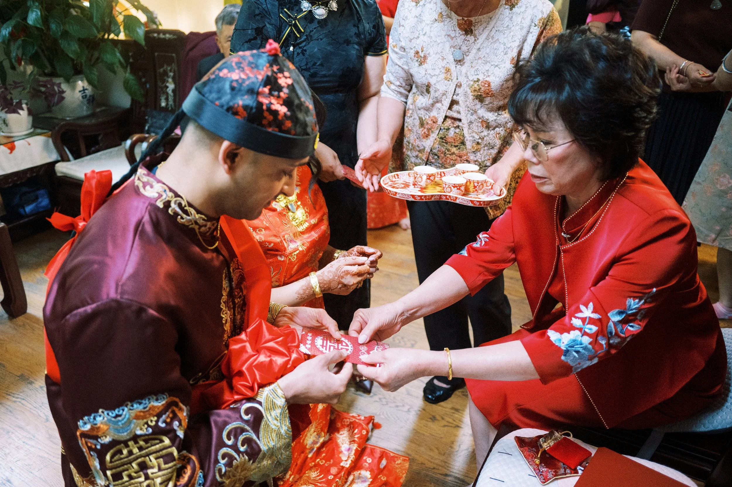 People participate in a traditional Chinese wedding tea ceremony indoors with decorations. A person in a floral dress and glasses serves tea to a man wearing a dark floral traditional Chinese wedding outfit and a cap. Others stand nearby, holding a r