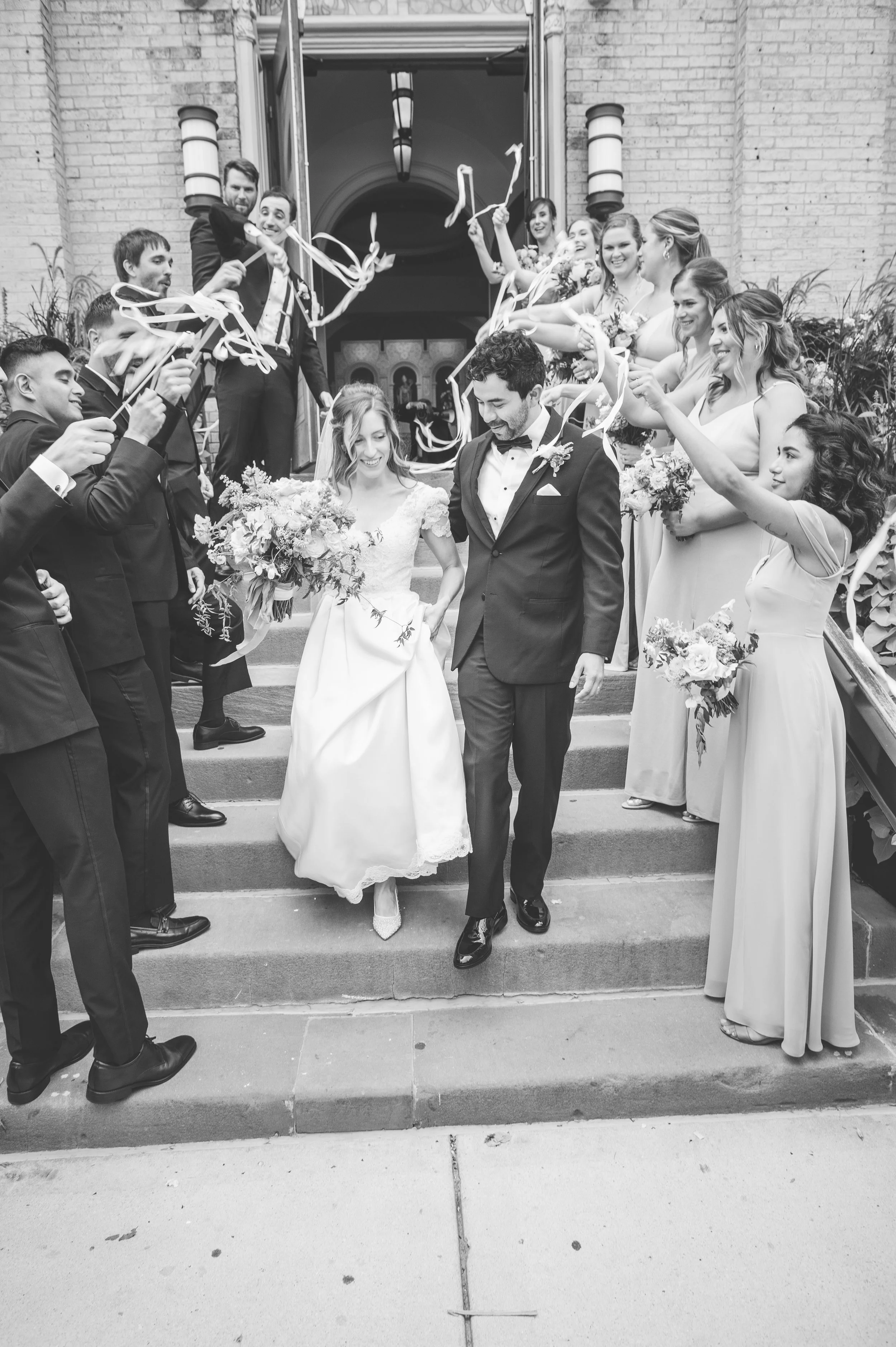 A group of smiling people celebrating a wedding on the steps of a building, with some holding ribbons and flowers.