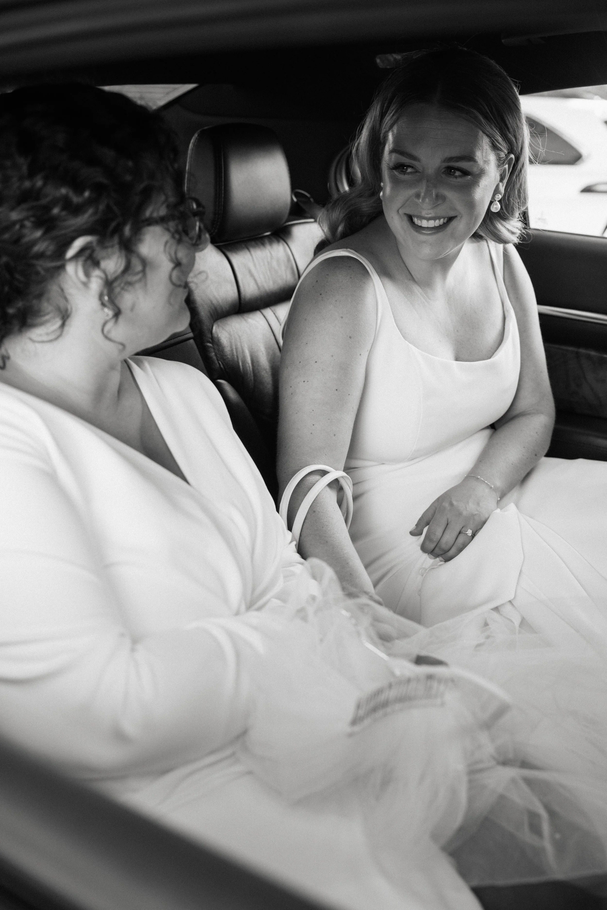 A woman in a white dress sitting in a car beside another woman, smiling and looking at her.