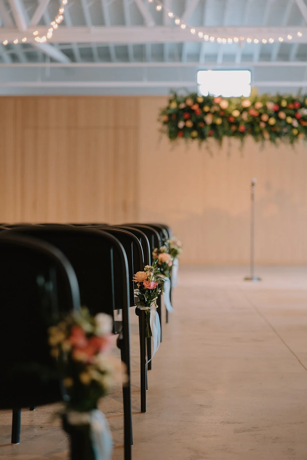 Empty indoor venue with black chairs decorated with small floral arrangements, a large floral centerpiece hanging from the ceiling, and a wooden wall background in a light-colored hall